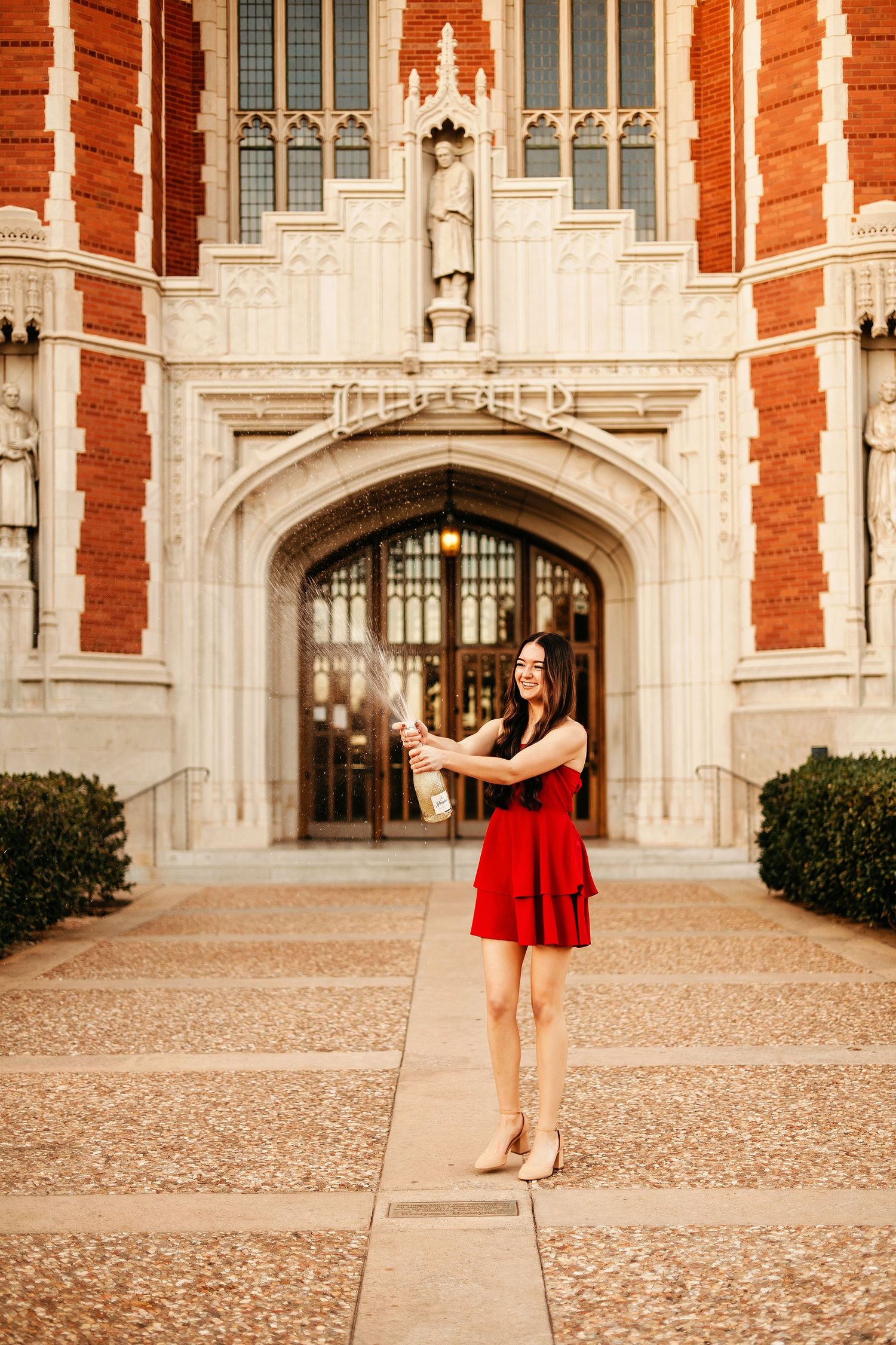 Woman in red dress popping champagne bottle in front of a grand brick university building entrance.