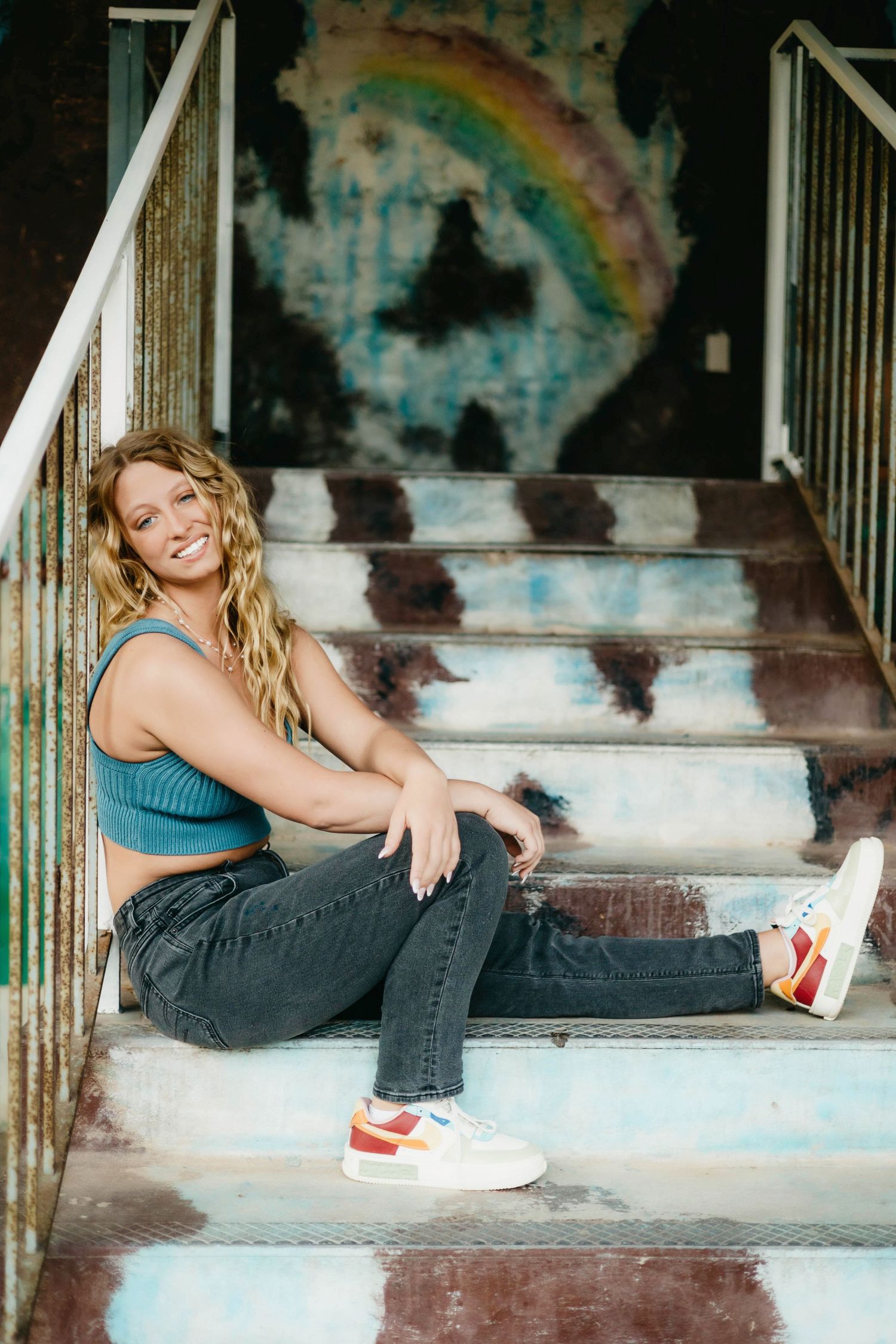 Smiling blonde woman in blue crop top and jeans sitting on rustic metal stairs with rainbow graffiti wall behind her.