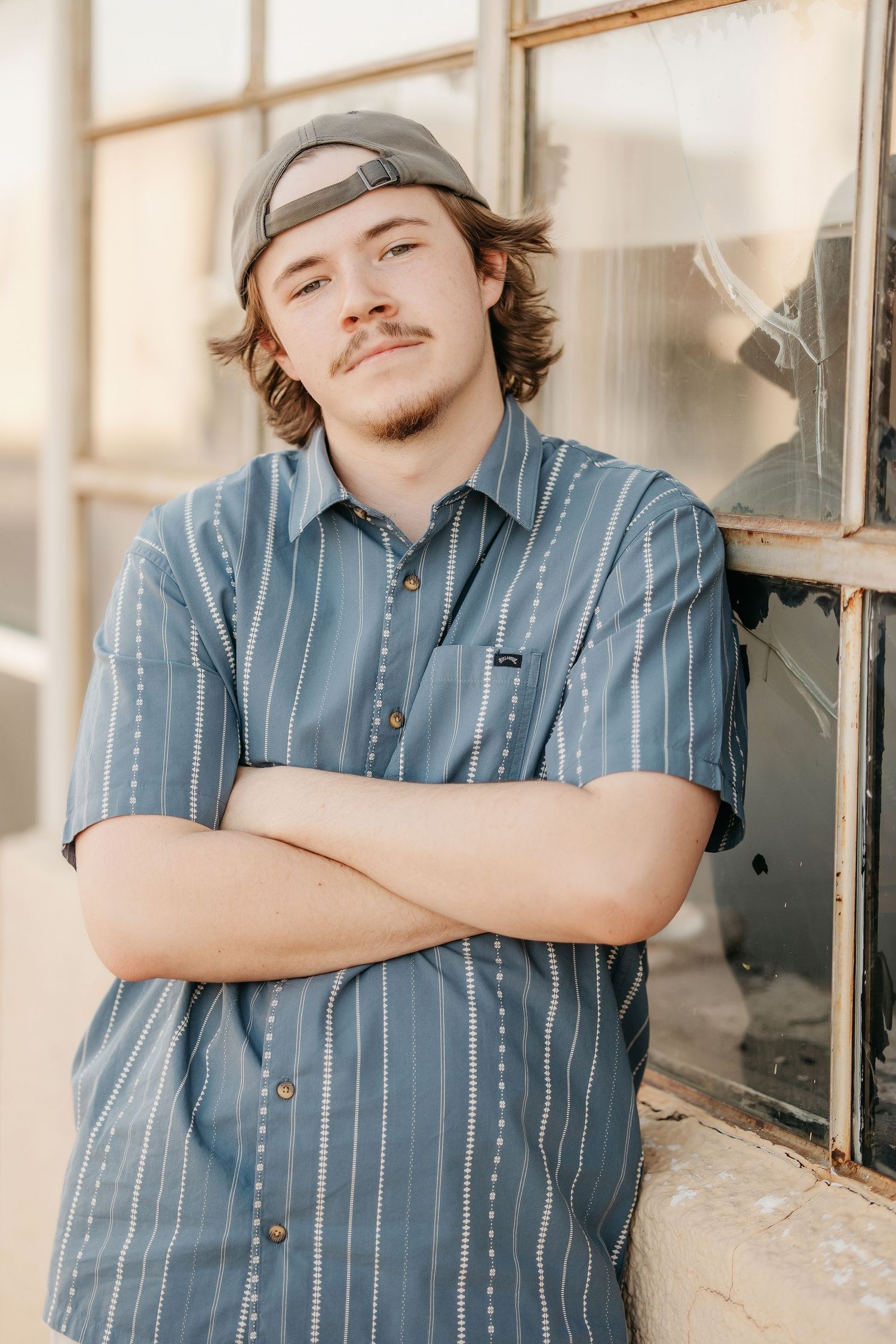 Young man with curly hair and snapback cap, arms crossed, wearing blue striped shirt, leaning against vintage window.