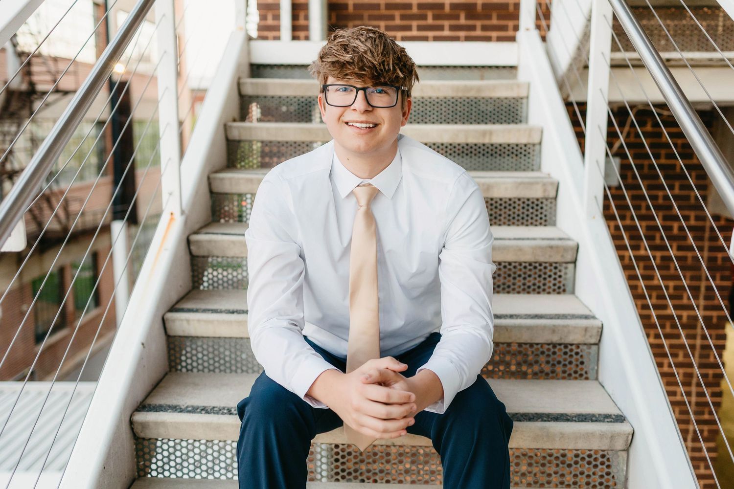 Young man in glasses, white shirt, and tan tie smiling on outdoor steps in a professional portrait.
