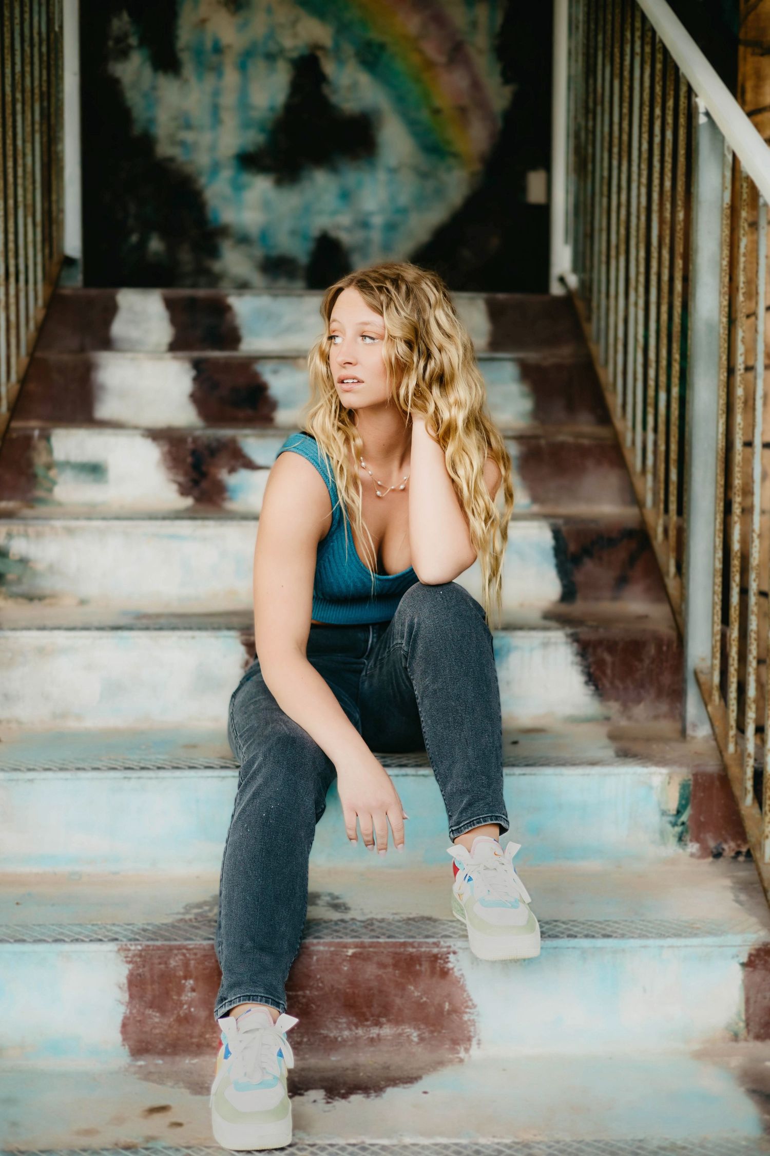 Young blonde woman sitting on weathered steps in teal top and jeans, posing against urban graffiti backdrop.