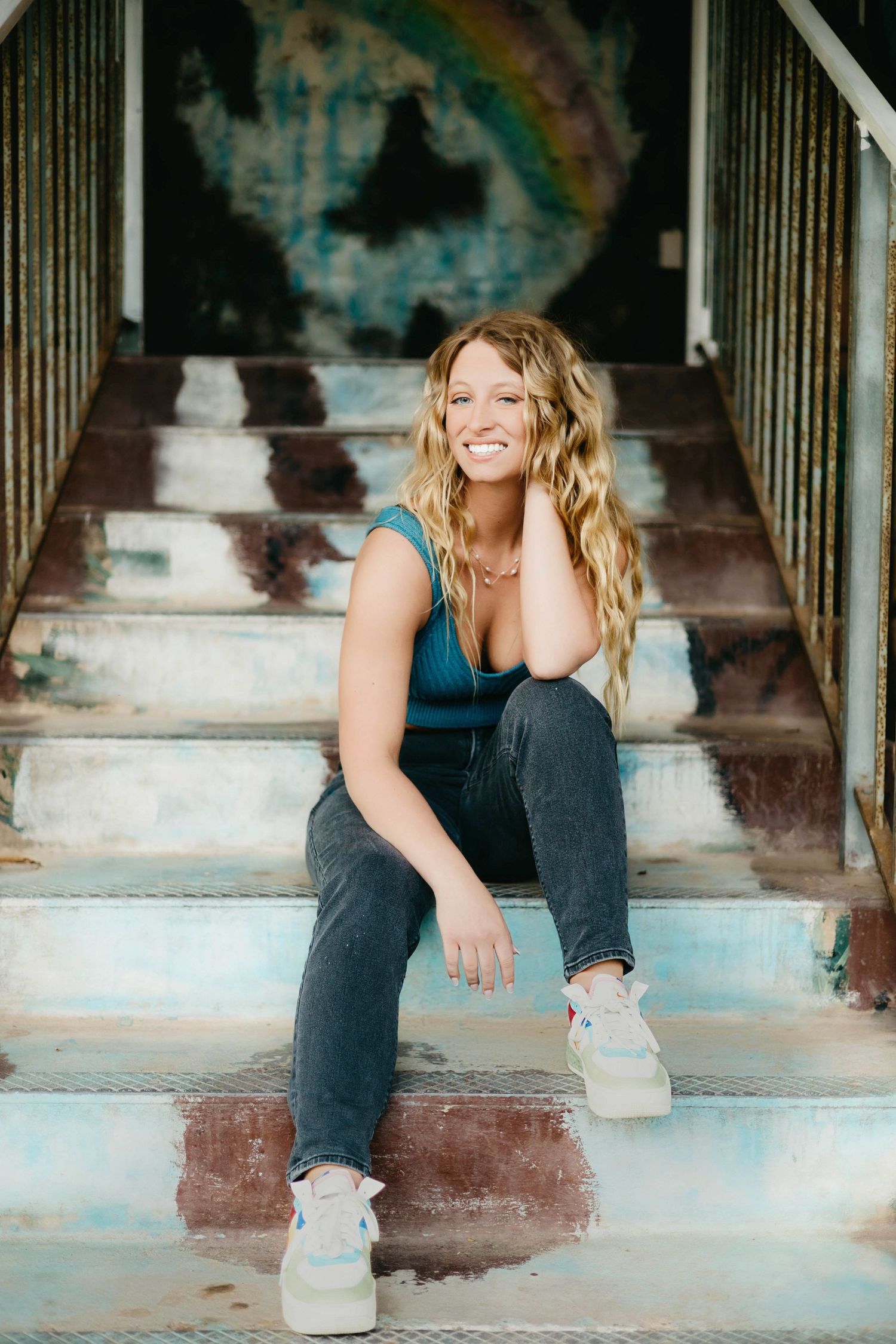 Smiling blonde woman sitting on worn steps in teal top and jeans, posing casually with hand on cheek outdoors.