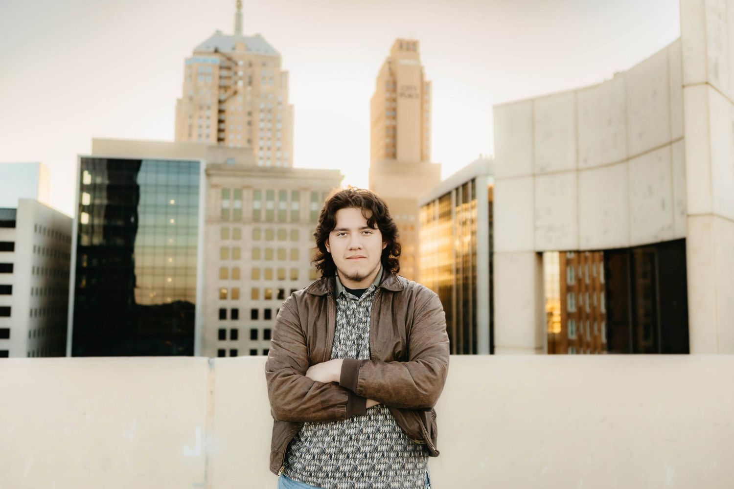 Young man with crossed arms standing on urban rooftop with city skyline and golden hour light behind him.