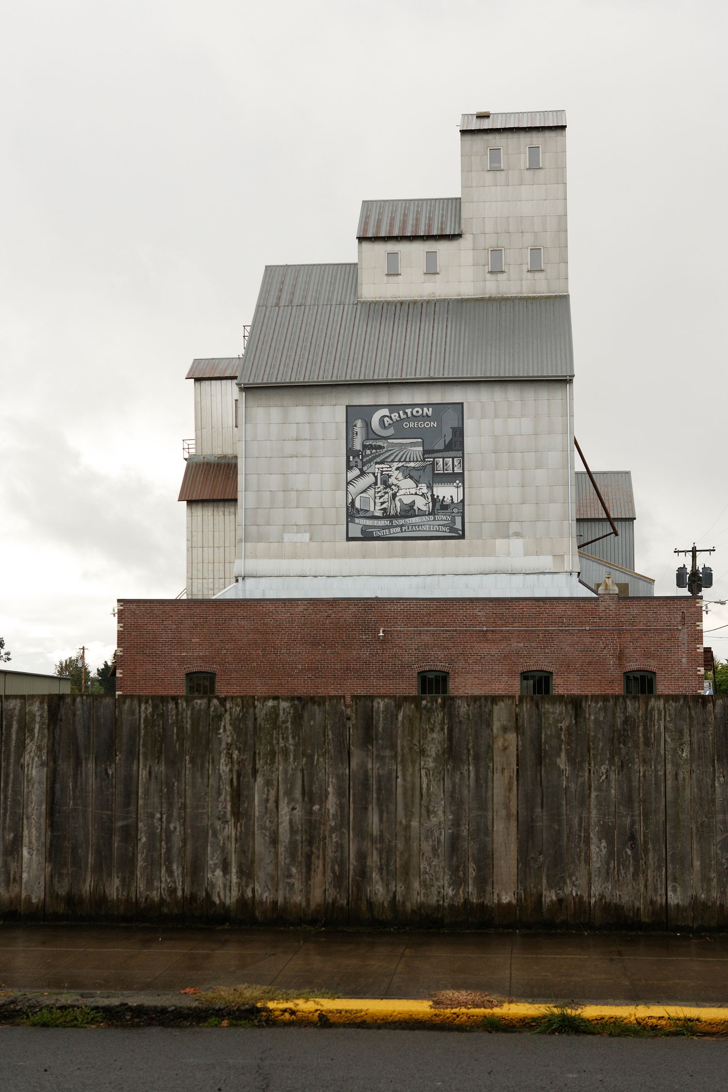 Old grain mill building with metal siding and wooden fence under overcast sky.