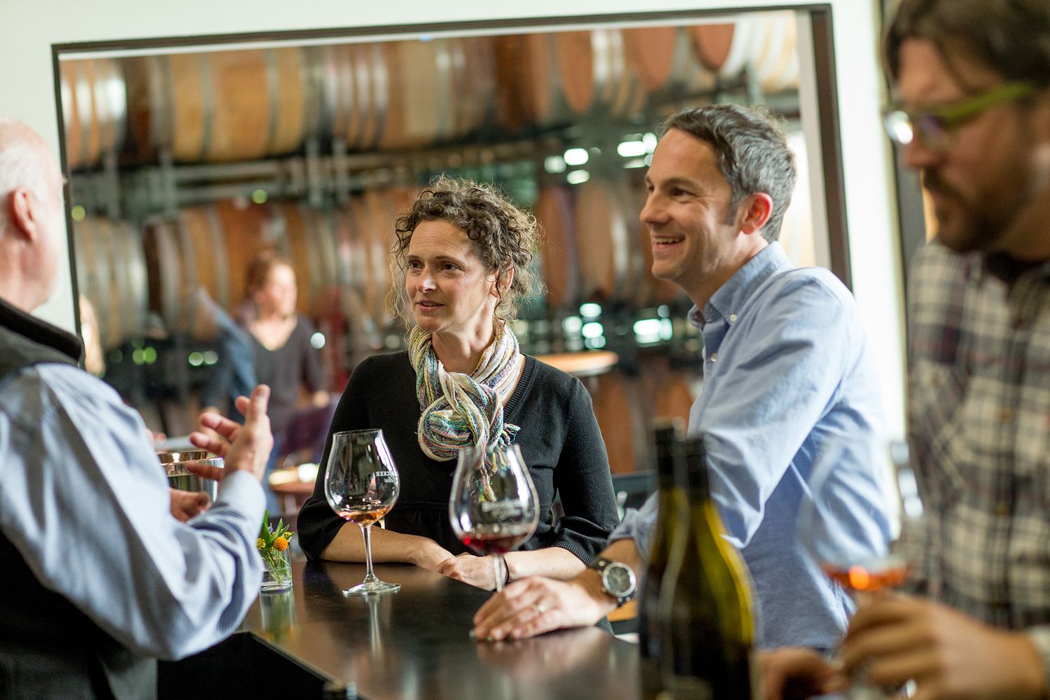 Group of adults enjoying wine tasting at a winery bar with wine glasses and barrels in the background.