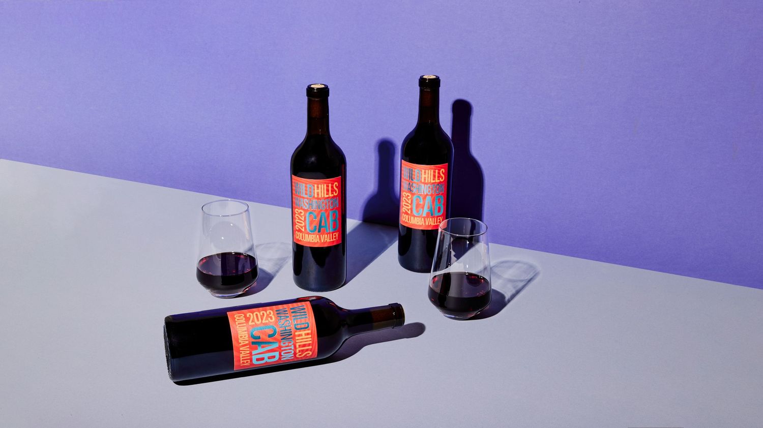 Three red wine bottles with orange labels and two glasses of red wine on a white surface against a bold purple background.