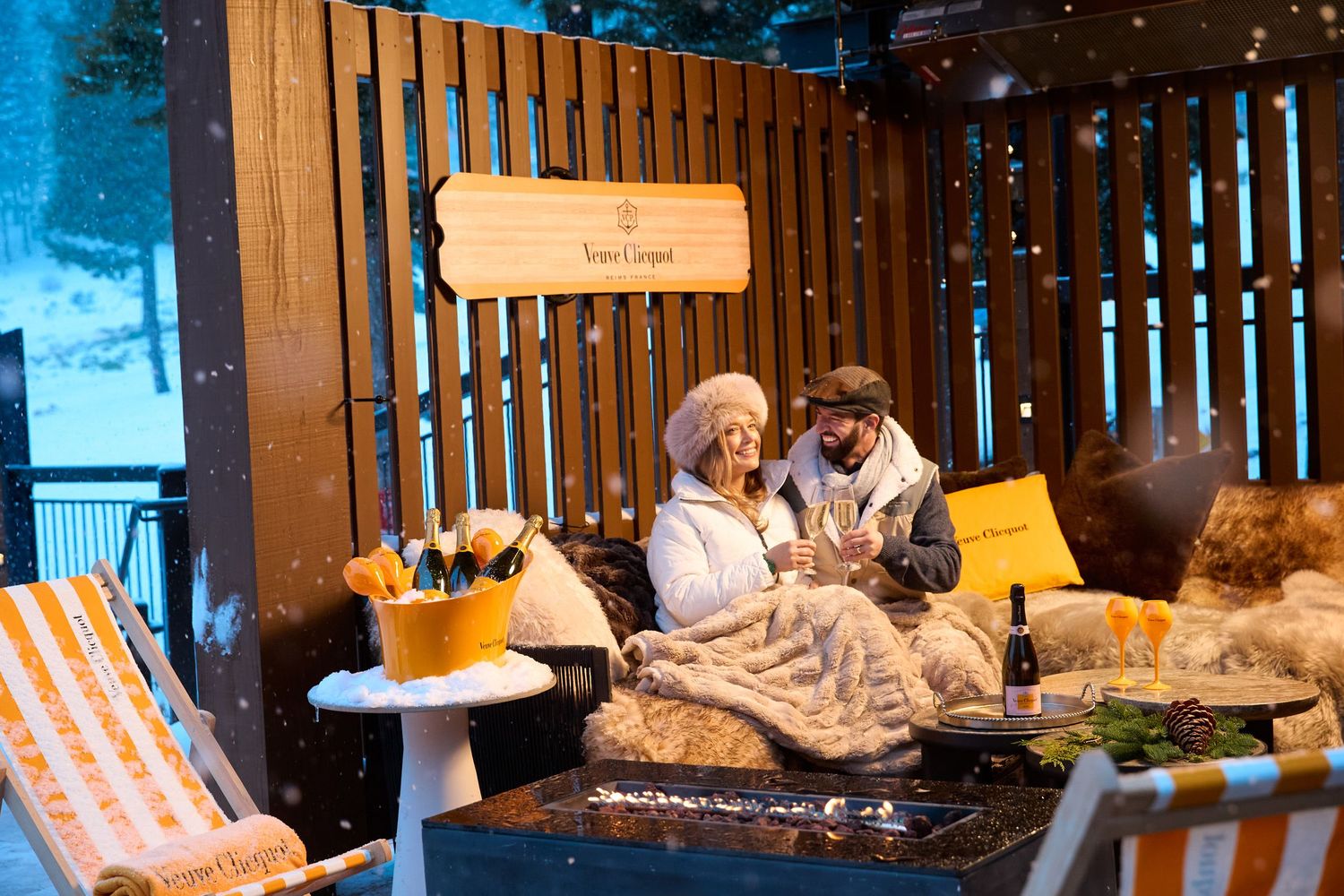 Cozy winter cabin interior with warm lighting and falling snow visible through large windows.