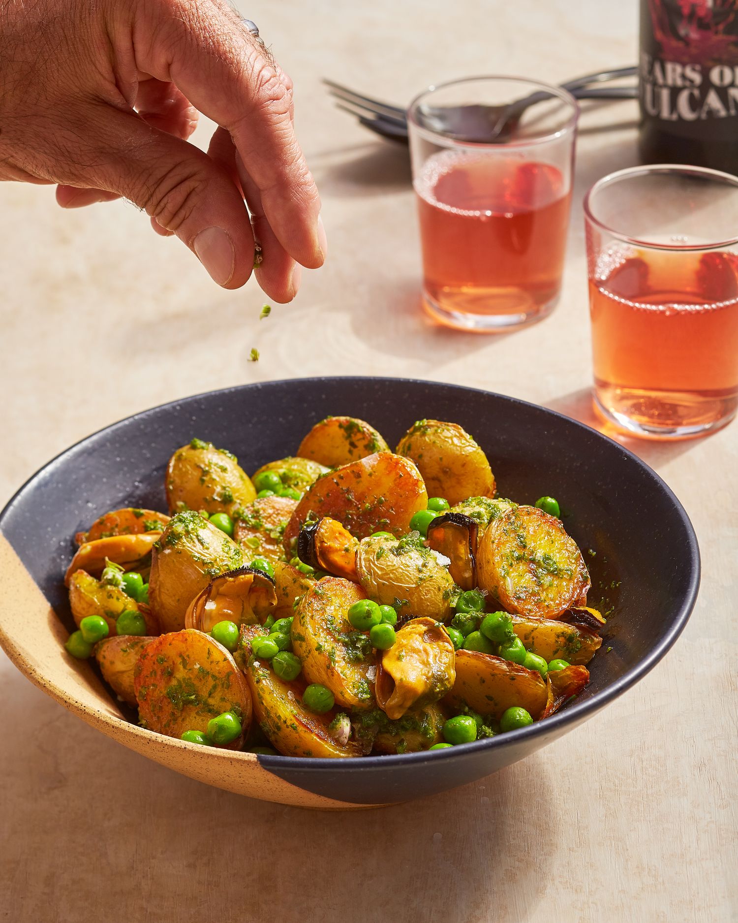 Roasted baby potatoes and peas in a blue bowl with pink cocktails in the background.