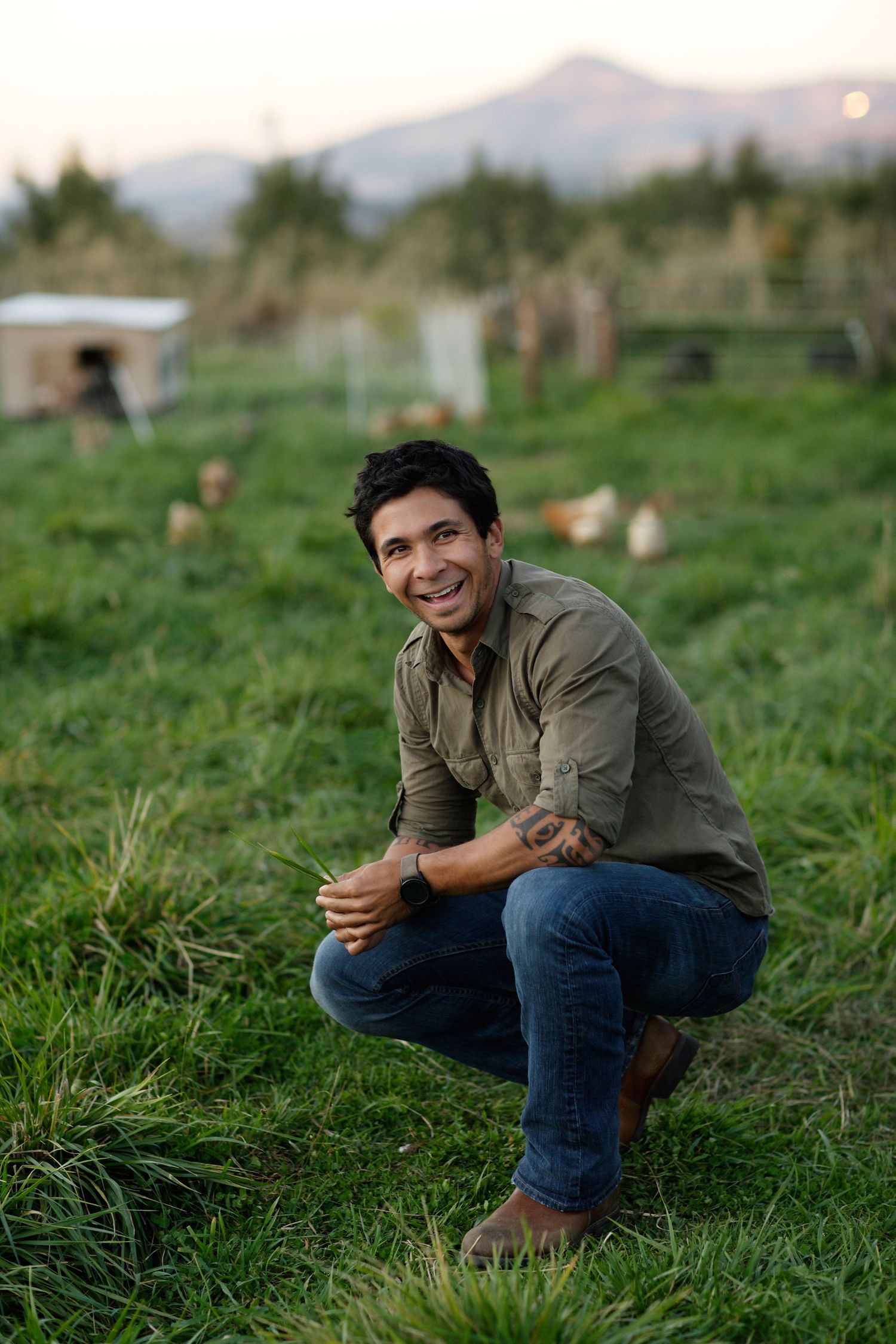 A person in casual attire crouches in a lush green field at sunset.
