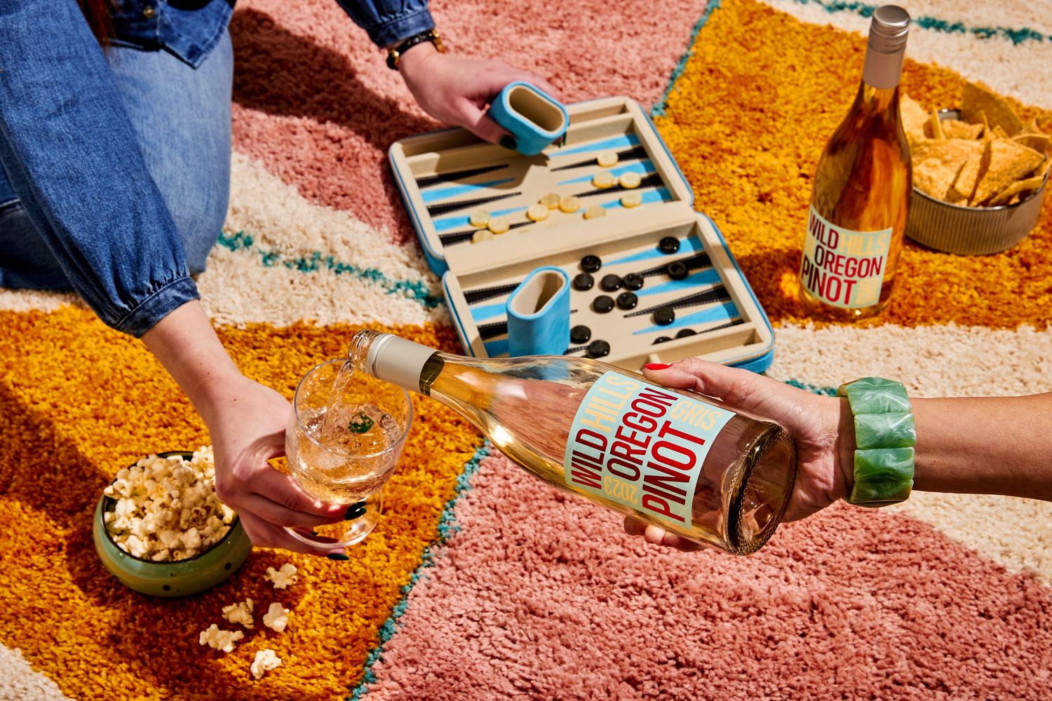 Two people pour Oregon Pinot wine into glasses beside a travel backgammon set, snack bowls and a second bottle on a colorful patterned rug.