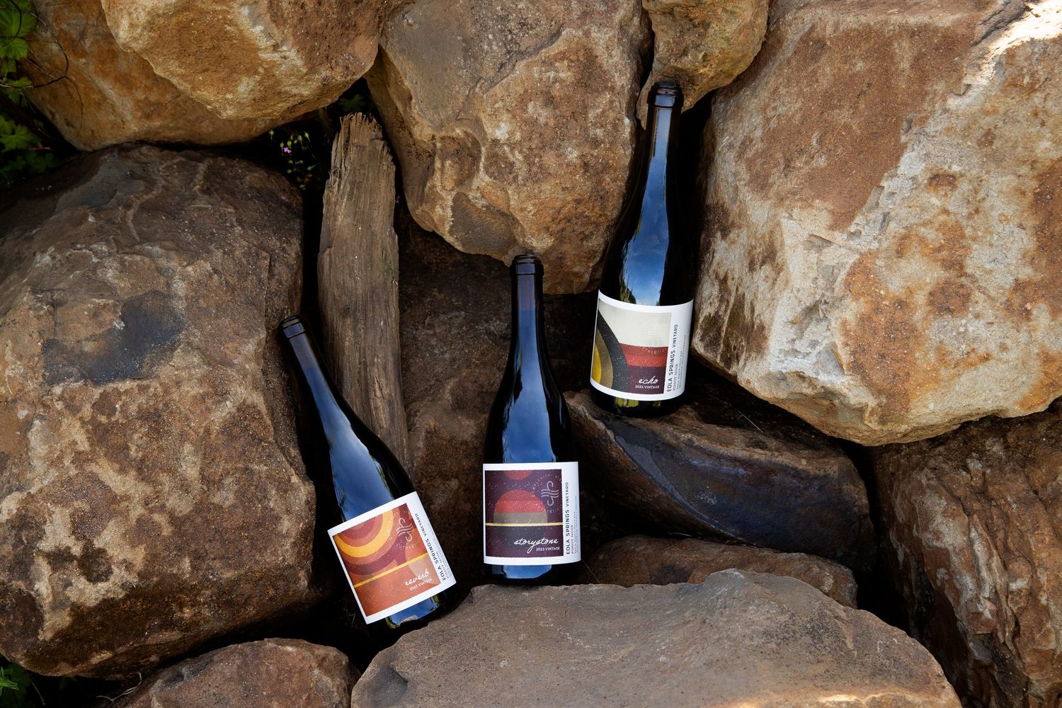 Three dark glass wine bottles with colorful labels rest at angles among large natural stones, creating a rustic vineyard-inspired display.