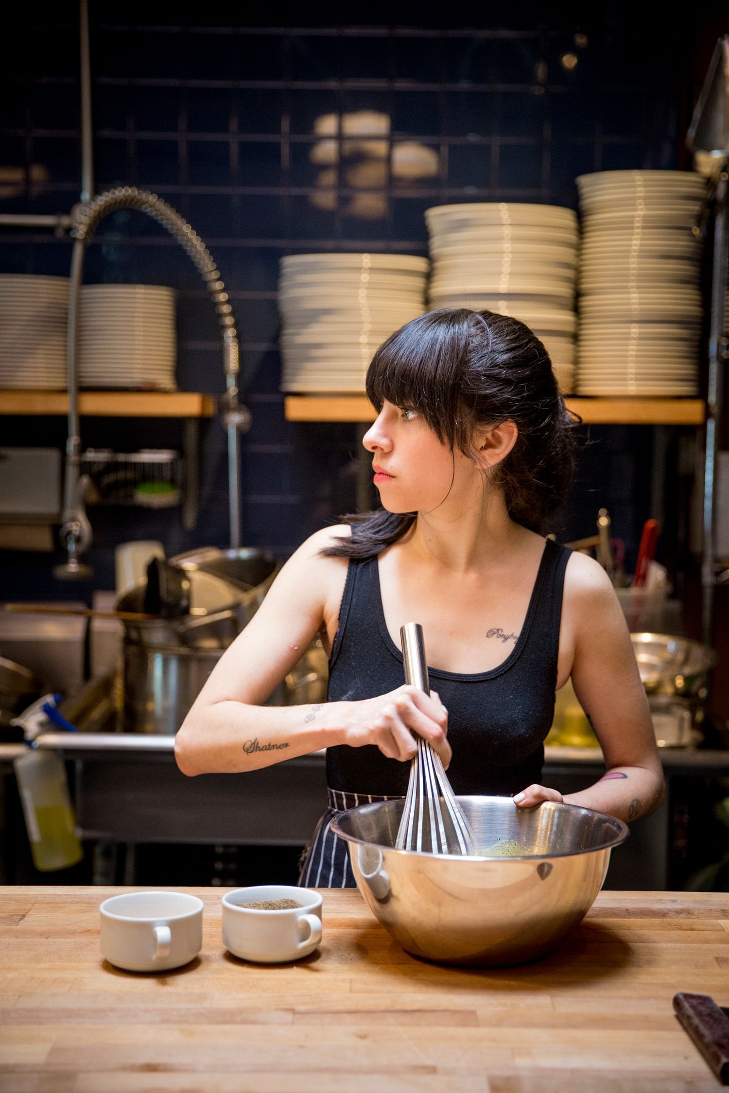 Chef whisking ingredients in a metal bowl in a professional kitchen with stacked plates.
