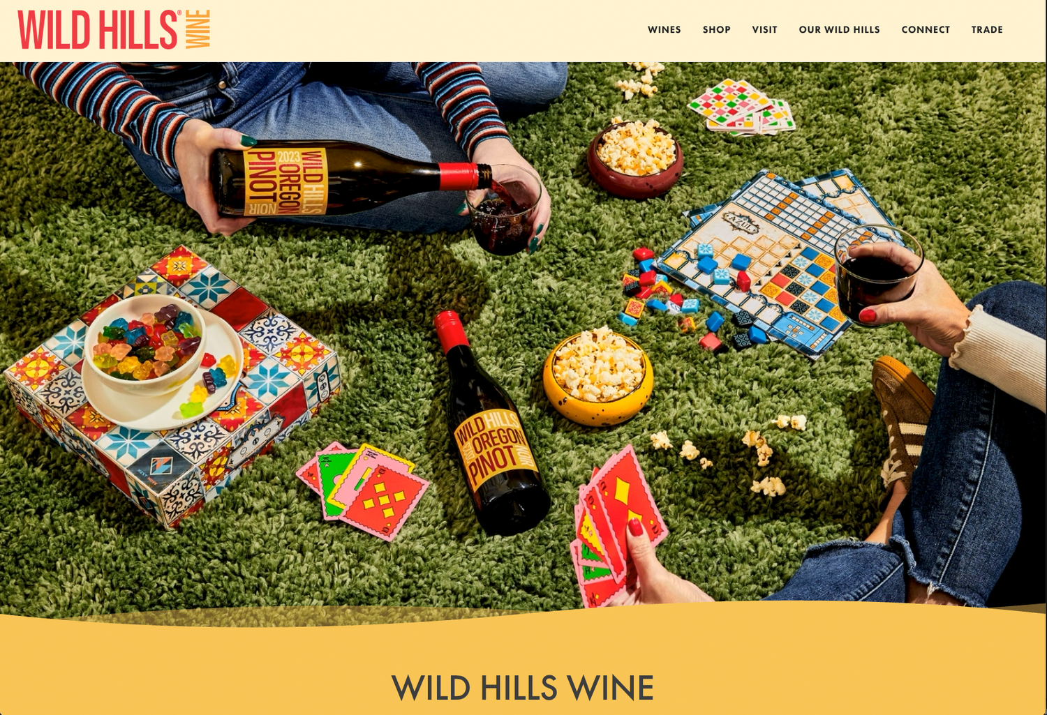 Wild Hills Wine website showing a picnic on grass with red wine bottles, glasses, snacks, colorful tile games and playing cards.