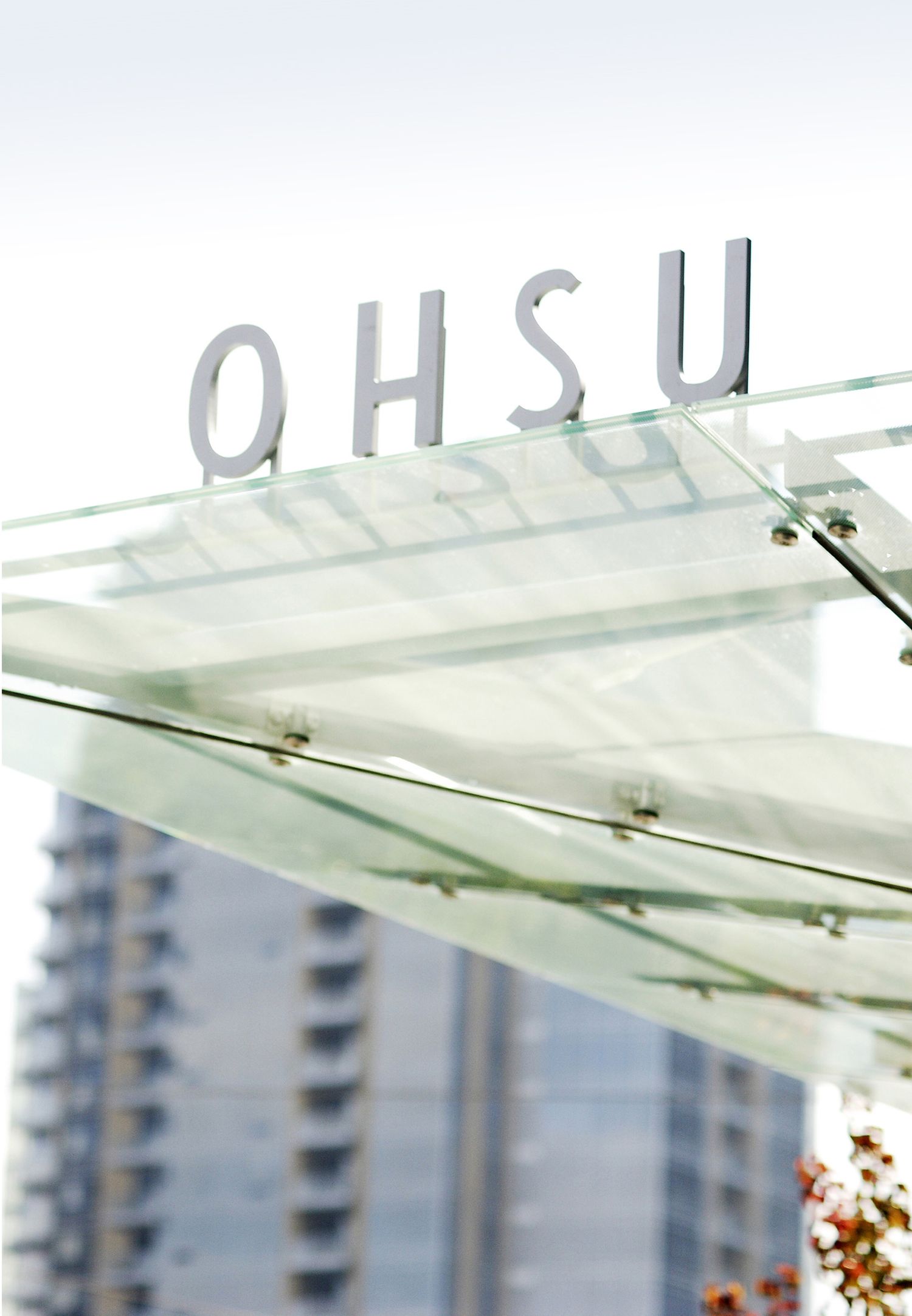 Letters spelling OHSU mounted on a glass structure with a tall building visible in the background.