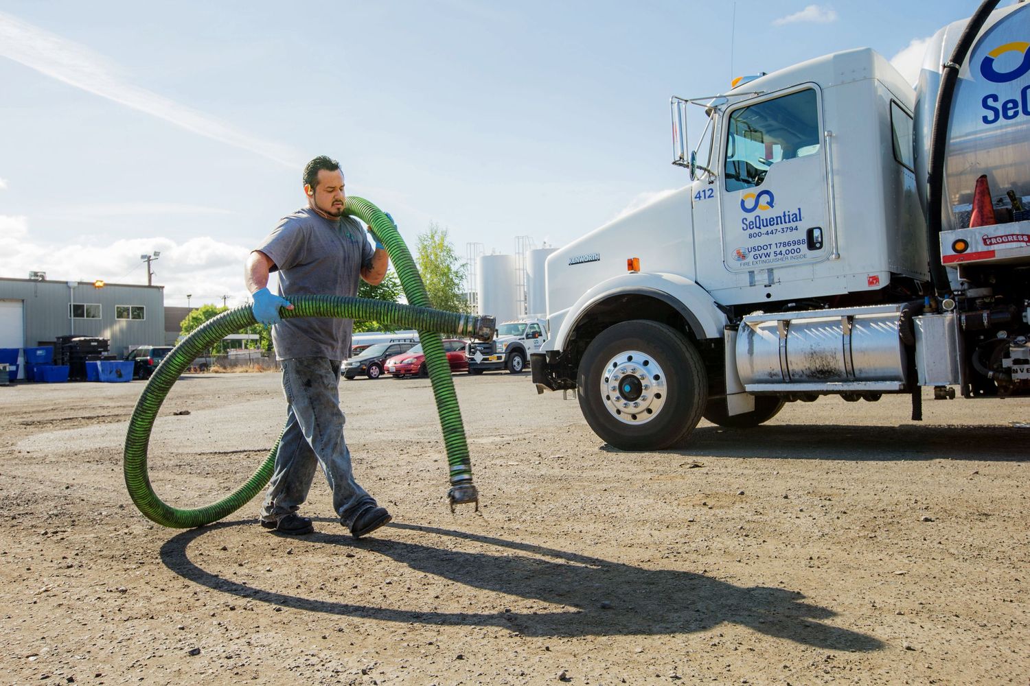 Worker operates large green hose near semi truck in industrial yard.