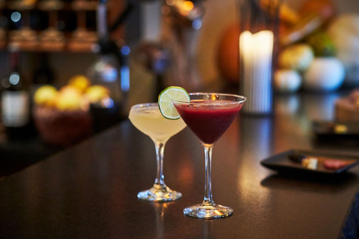 Two elegant cocktails on a dark bar counter, one red and one pale yellow with lime garnish.