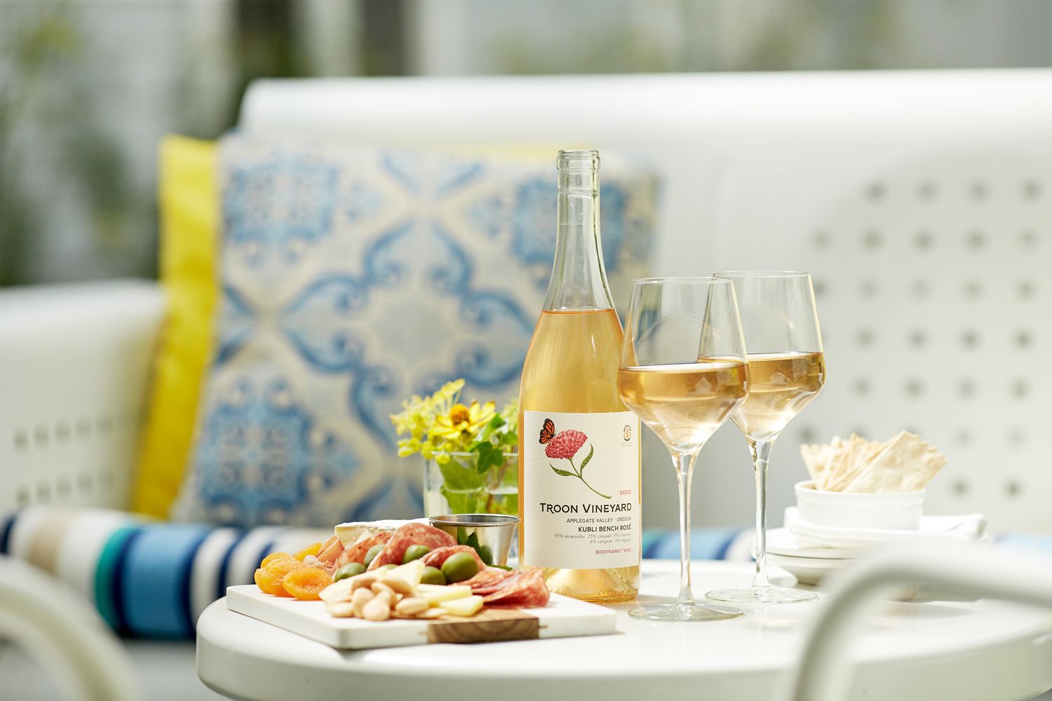 A bottle of Troon Vineyard wine with three glasses and charcuterie on a white outdoor patio table.