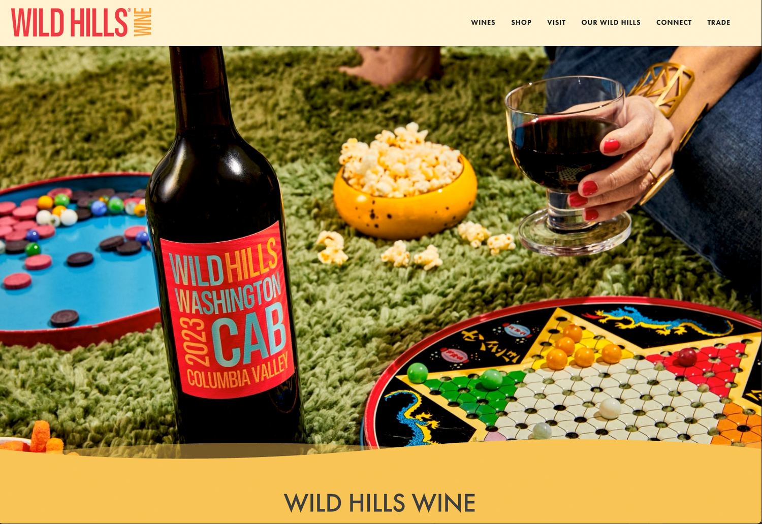 Wild Hills Washington Cab bottle beside a glass of red wine, popcorn bowl and colorful game boards on a green rug.
