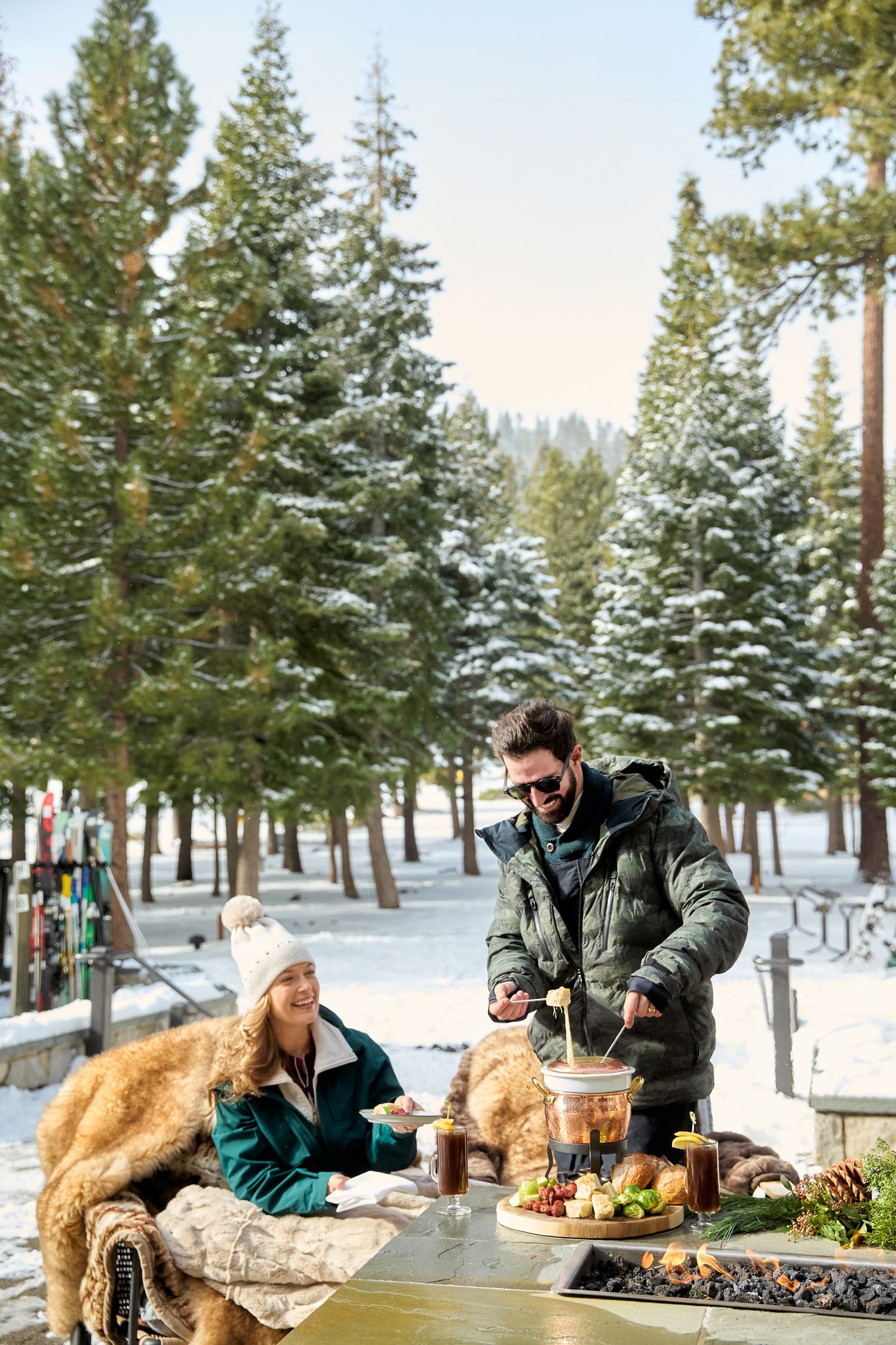 People enjoying a winter picnic with food and drinks at an outdoor table surrounded by snow-covered pine trees.