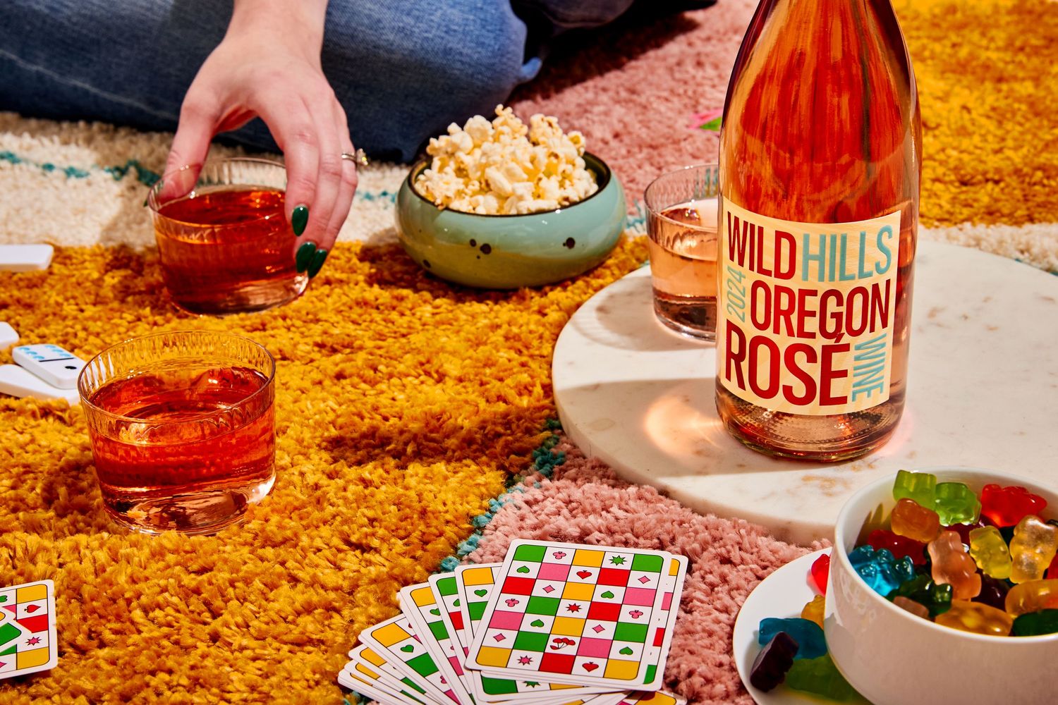 Bottle of Wild Hills Oregon Rosé on a table with glasses of wine, popcorn, candy and playing cards on a shaggy mustard rug.