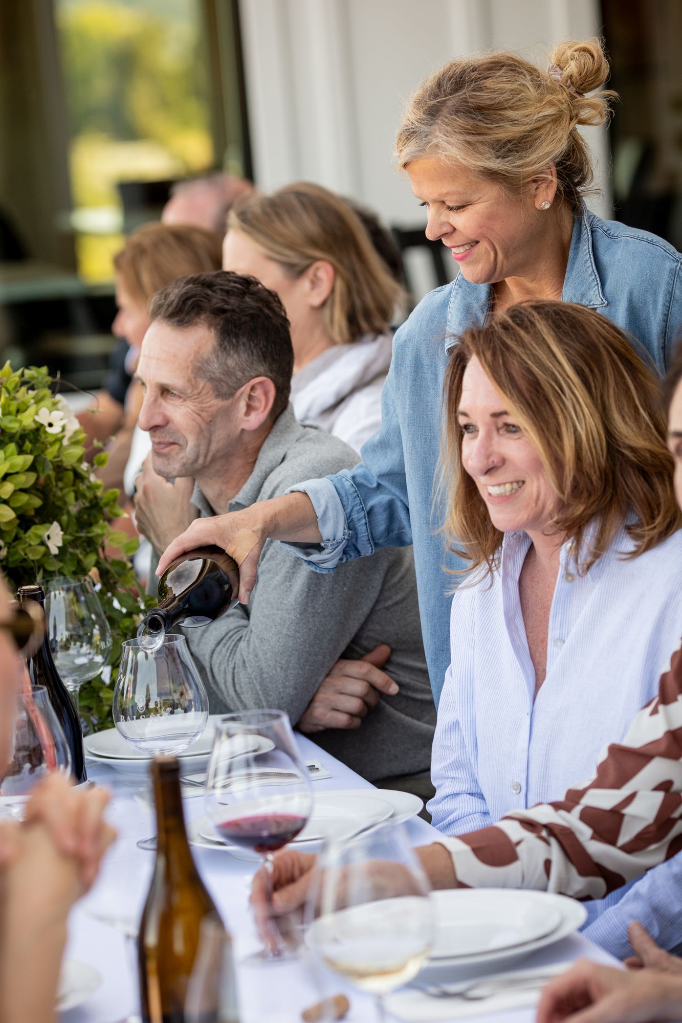 Group of adults enjoying a wine tasting dinner party outdoors, with a woman pouring red wine at a festive table.