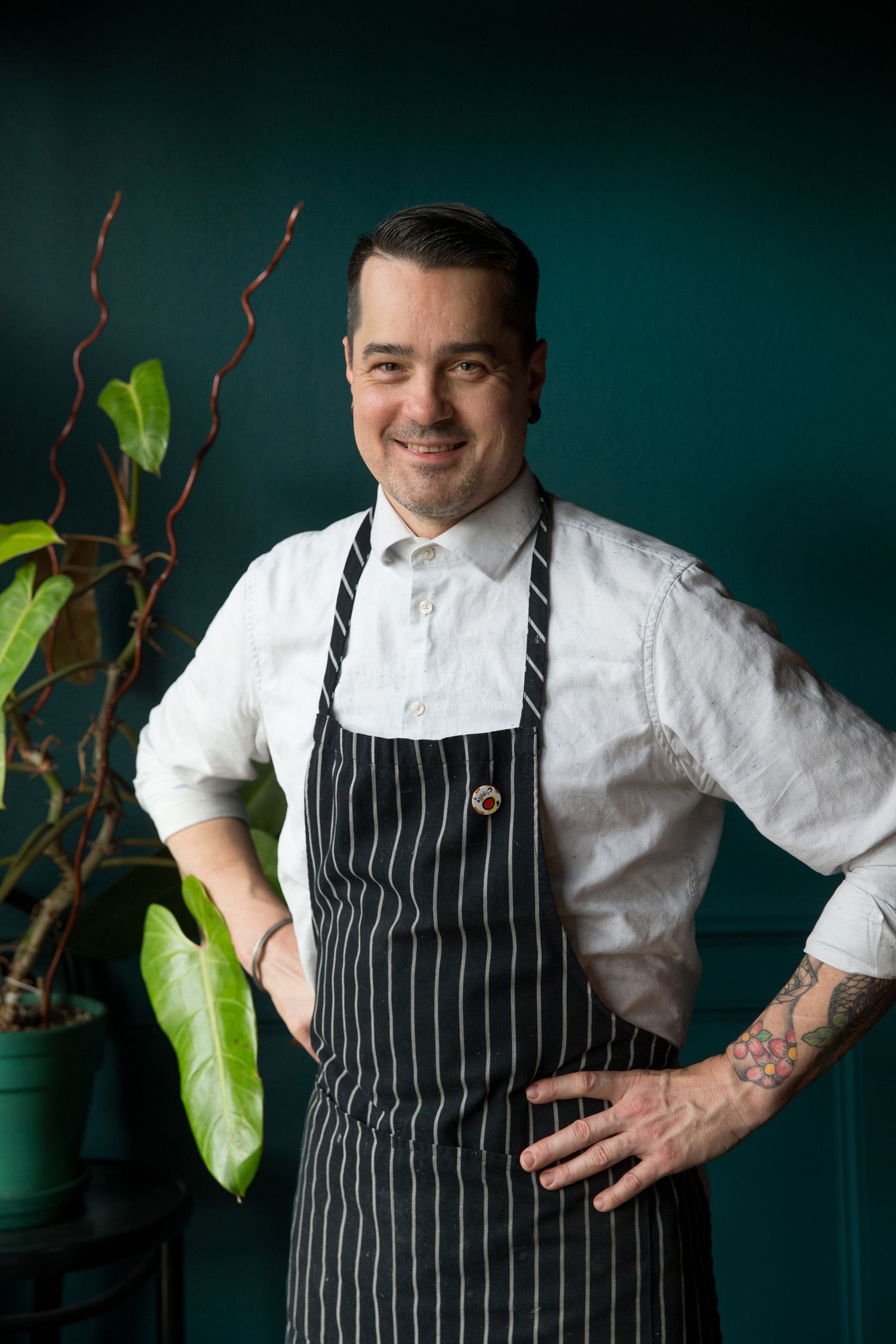 Professional in striped apron and white shirt standing against dark teal wall with decorative plants.