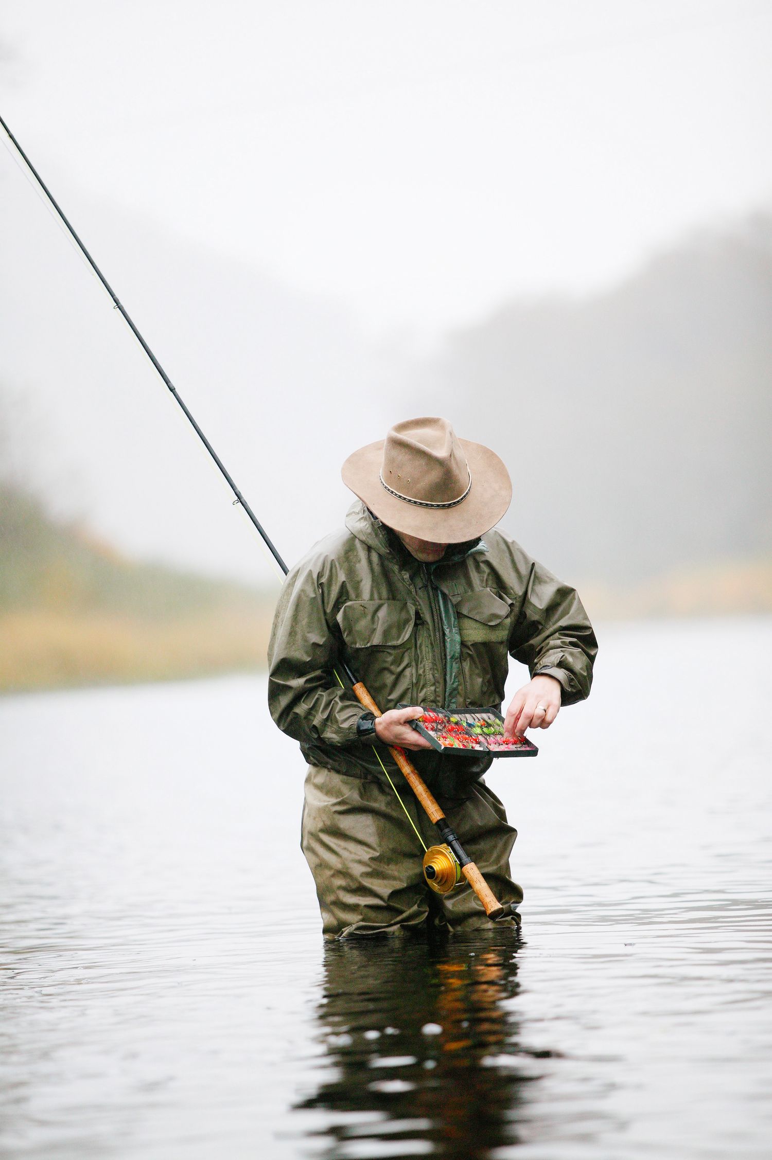 A fly fisherman wearing waders and a hat stands in a misty river while fishing.