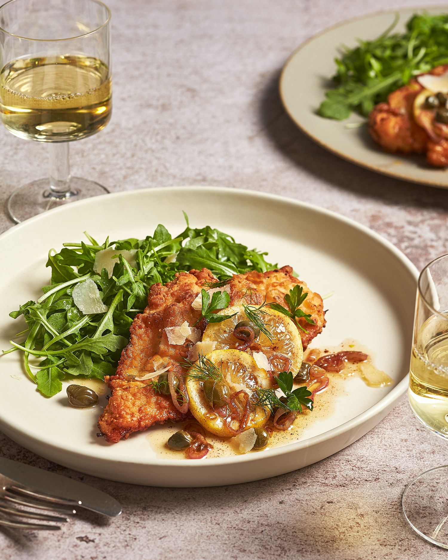 Breaded chicken cutlet garnished with fresh herbs and lemon on white plate.
