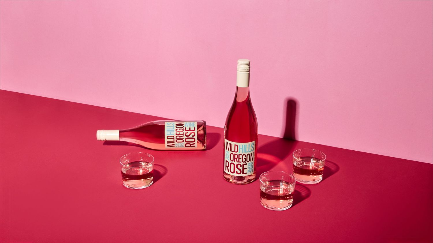 Two bottles of Wild Oregon Rosé, one standing and one lying down, beside three glasses of rosé wine on a bold pink and magenta backdrop.