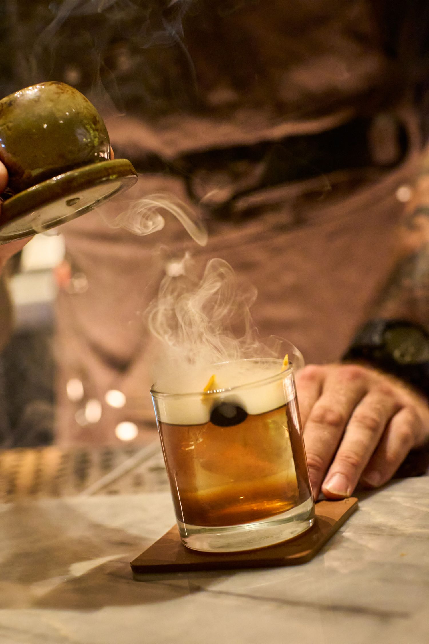 A hand pours amber-colored whiskey from a bottle into a lowball glass on a dimly lit bar counter.