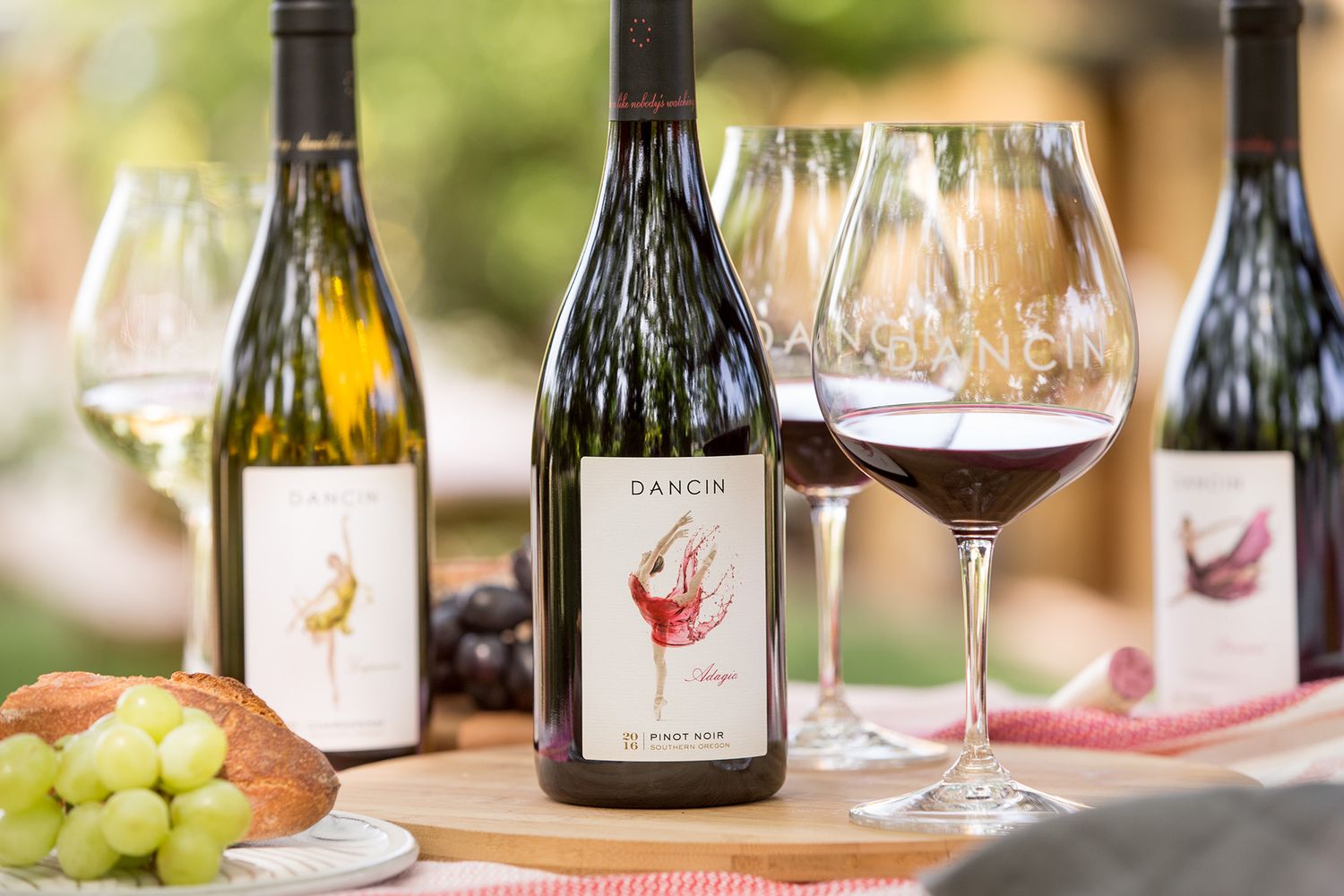 Dancin Vineyards wine bottles and glasses on a wooden board with green grapes and bread in an outdoor setting.