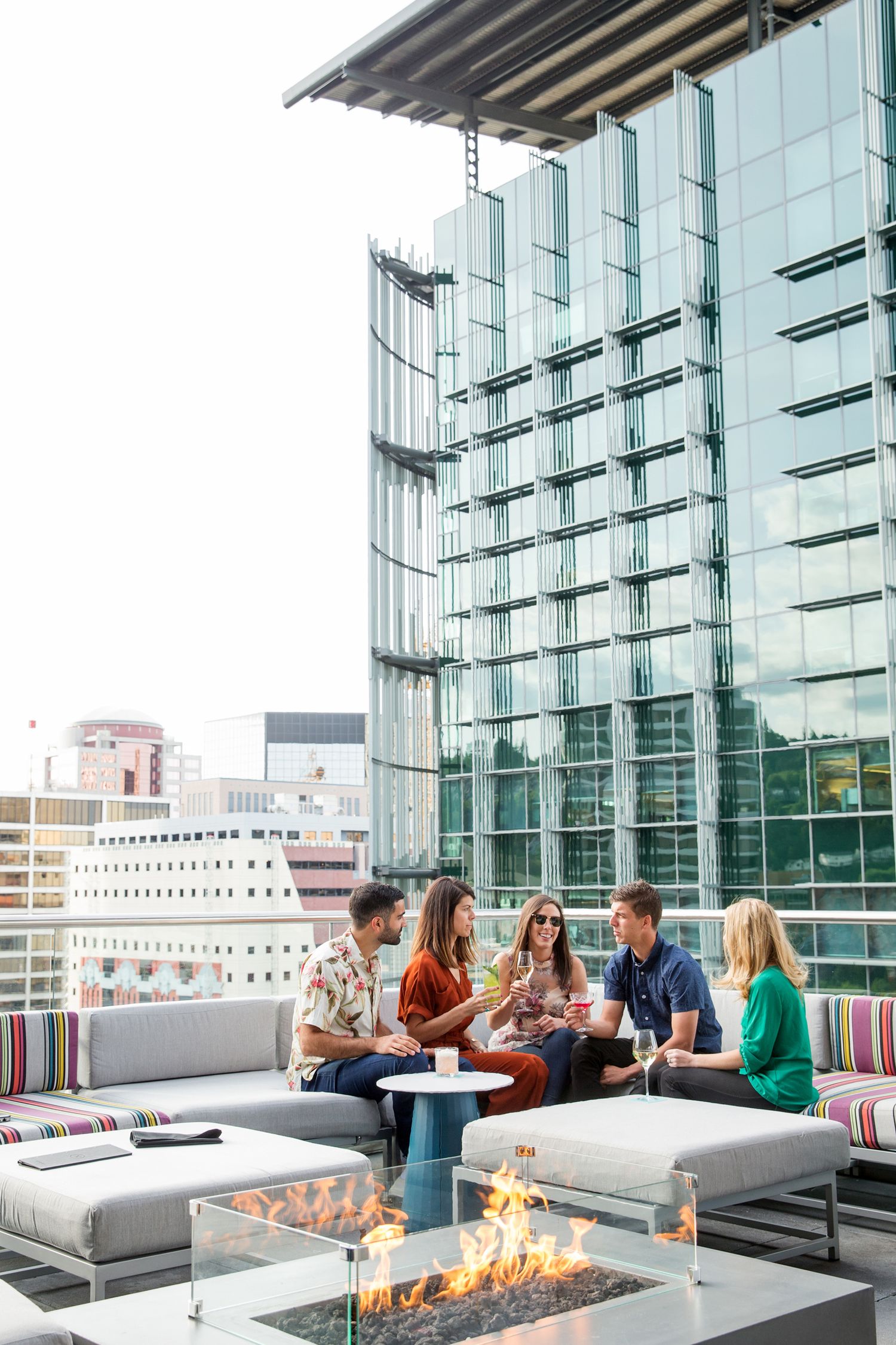 Modern rooftop lounge with glass walls, grey seating, and fire pit overlooking city skyline.