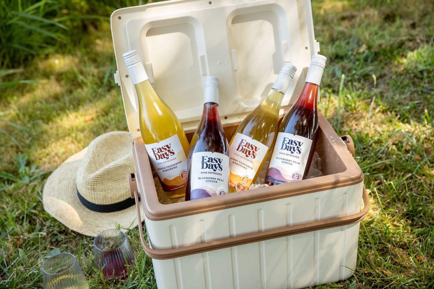 Open cream cooler holding four bottles of white, rosé and red wine on a sunny lawn next to a straw hat and glasses.
