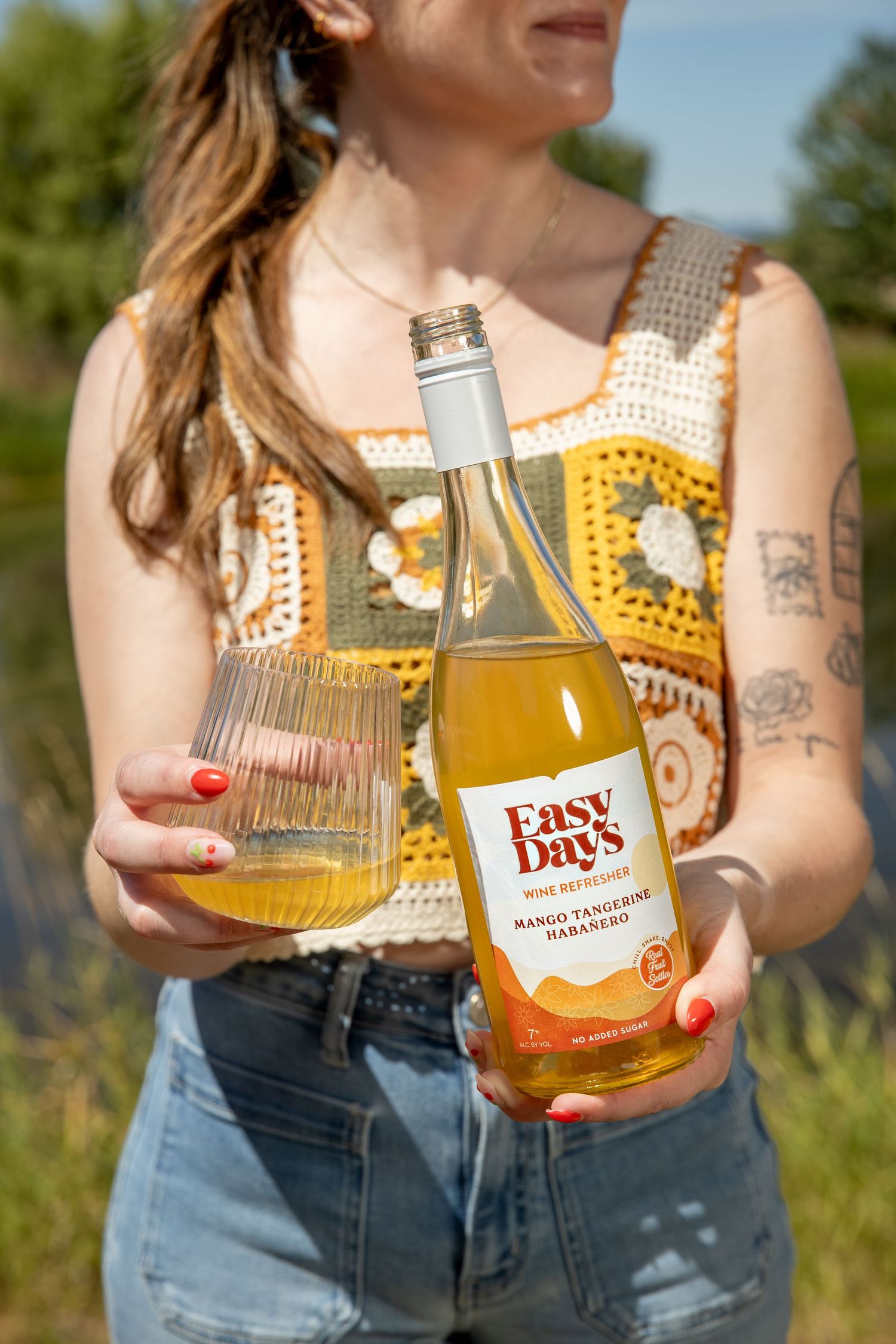 Woman in a crochet top holds a bottle of Easy Days Mango Tangerine Habanero wine refresher and a filled glass outdoors.