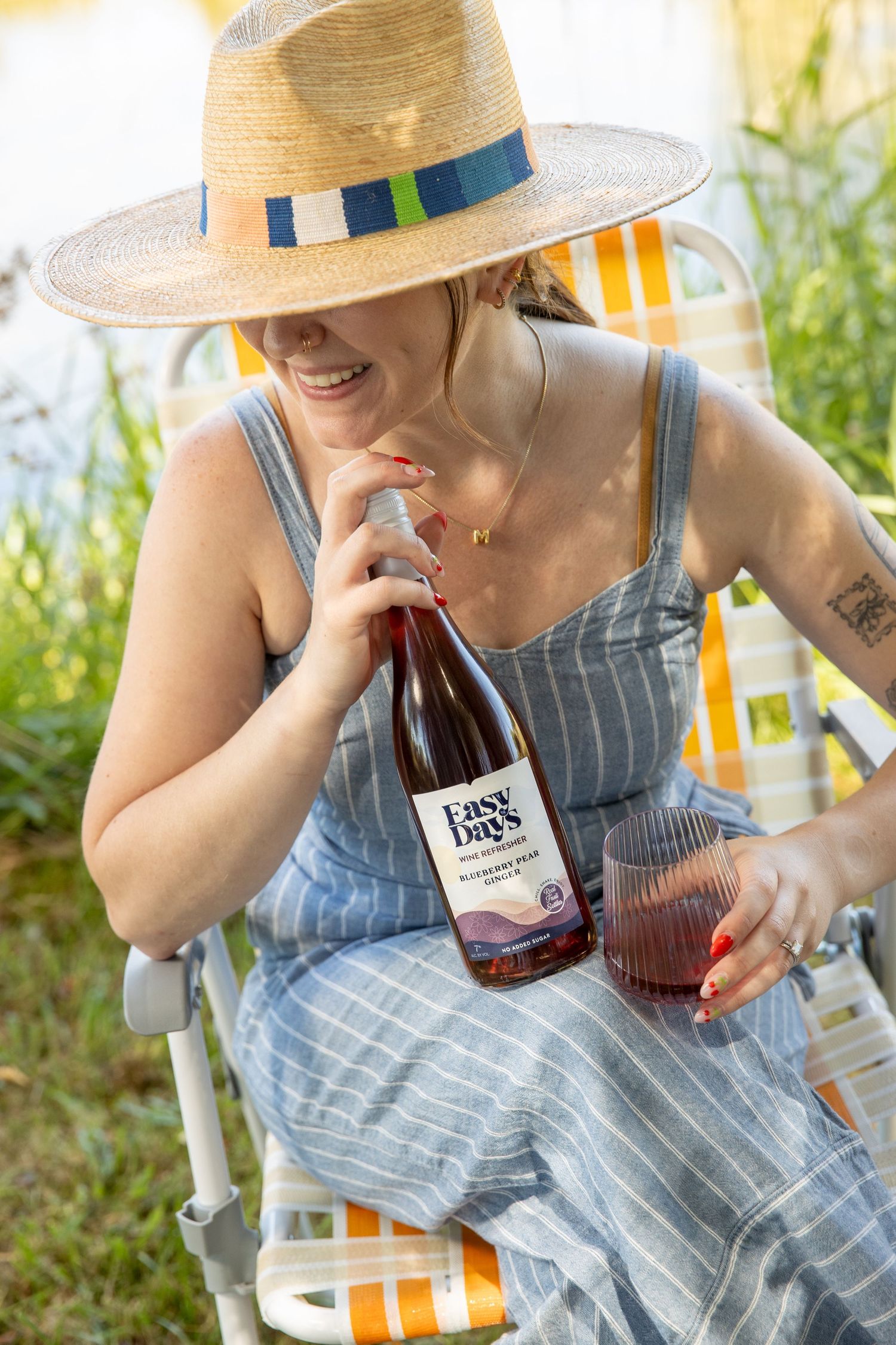 Smiling woman in a straw hat and striped dress sits in a lawn chair by the water, holding a bottle of Easy Day's rosé and a glass of wine.
