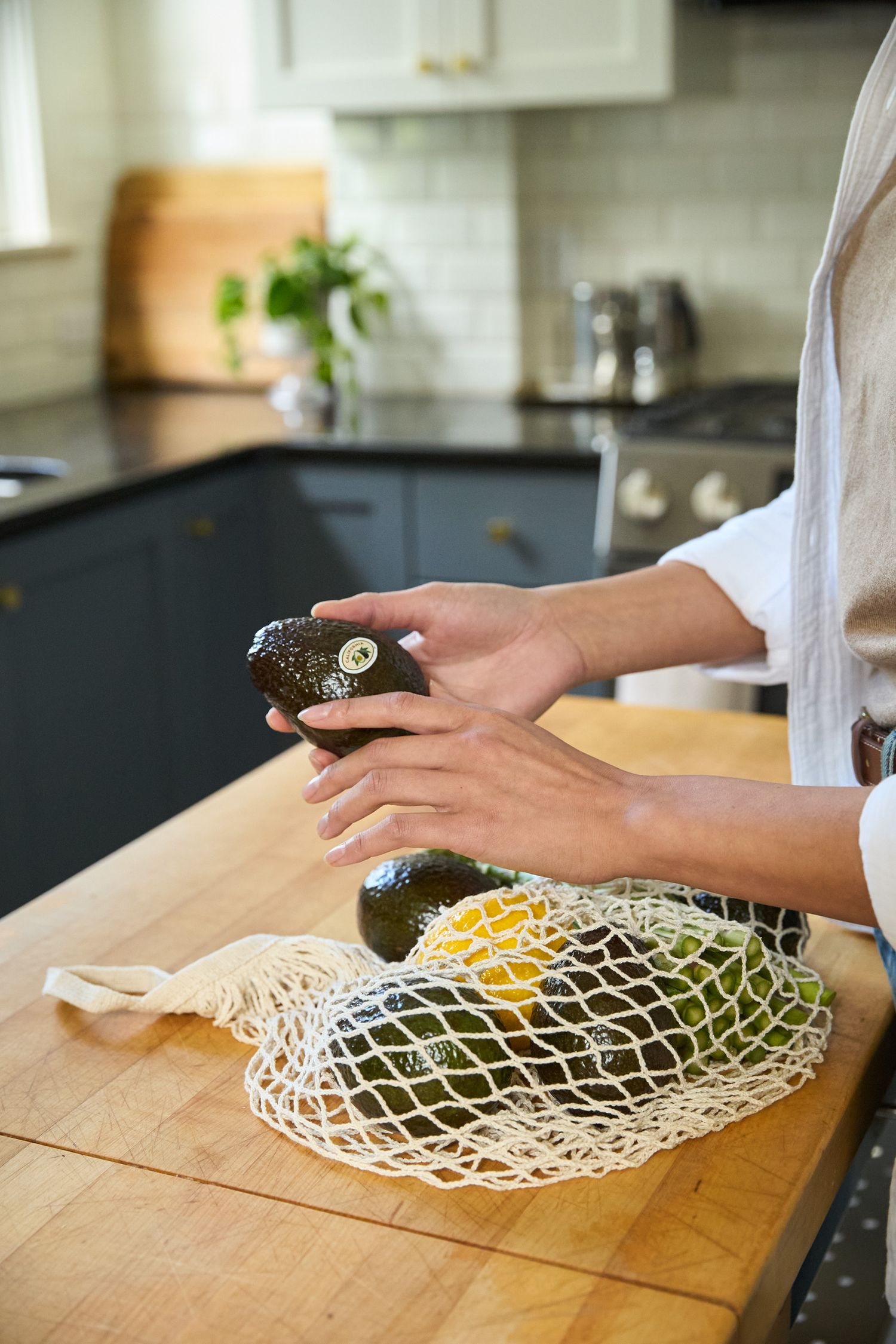 Hands place fresh avocados into a white mesh produce bag on a wooden cutting surface.