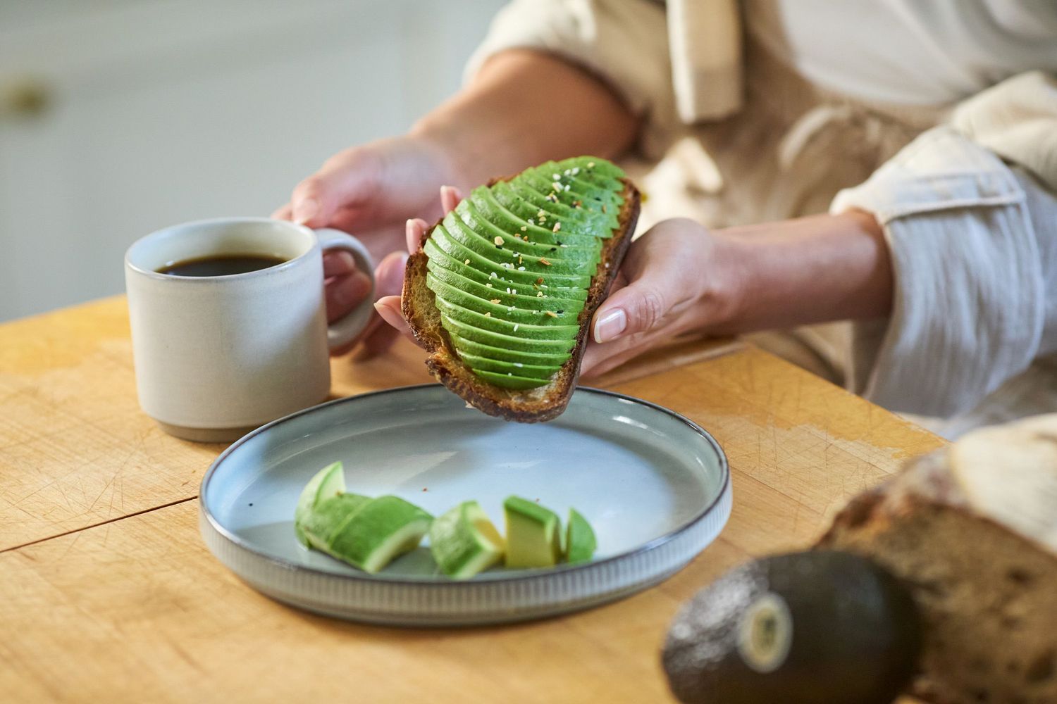 Someone spreading fresh avocado on toast with a coffee cup and plate on wooden table.