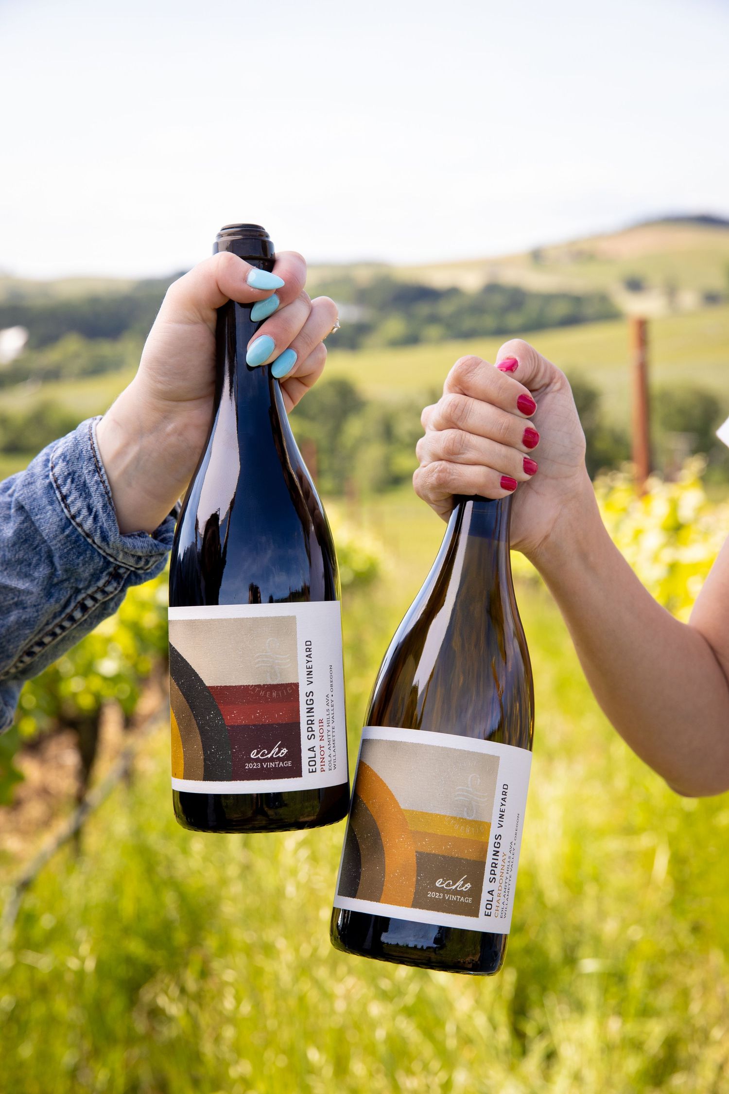 Two hands with painted nails clink Eola Springs Vineyard wine bottles together in a sunny, green vineyard with rolling hills.