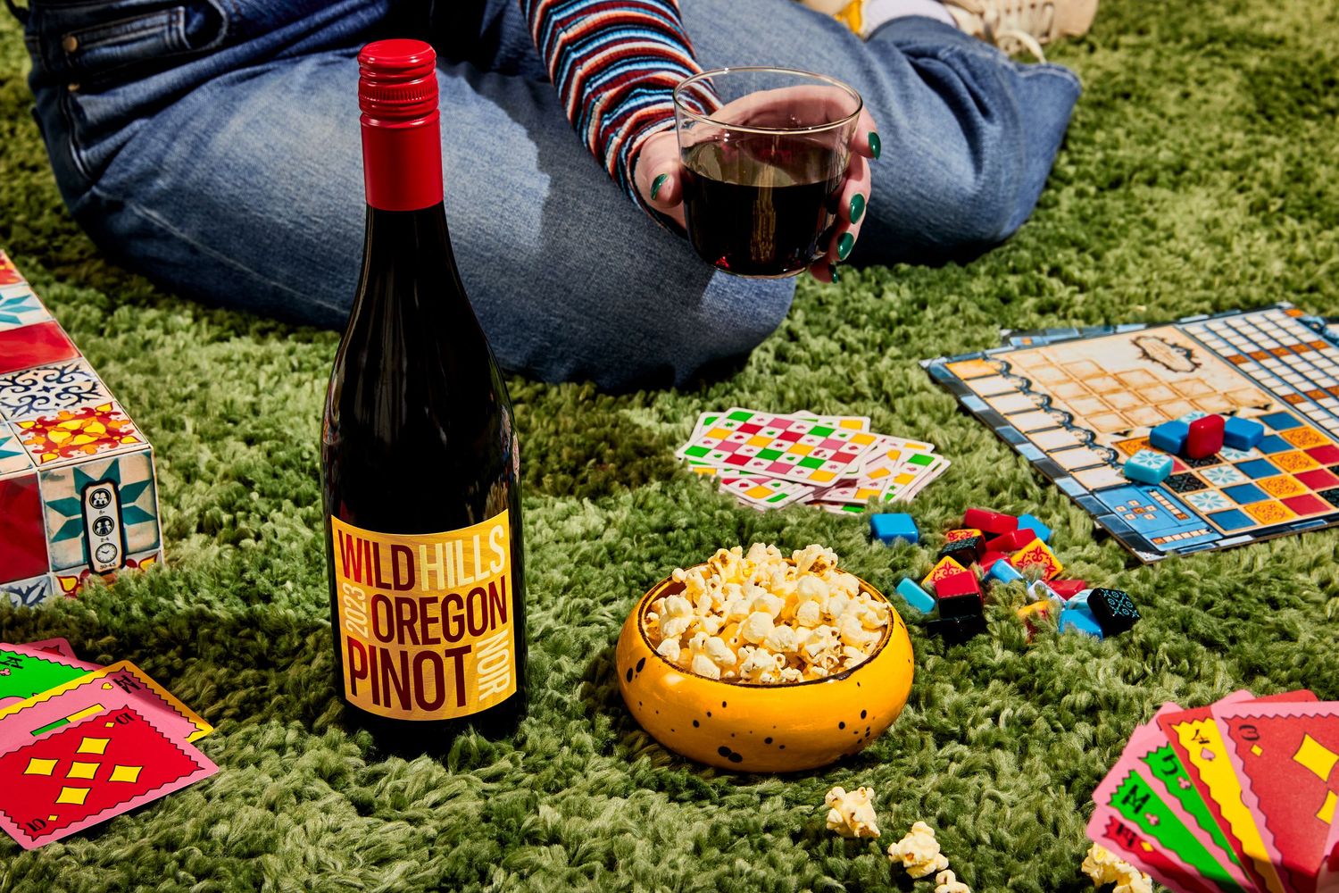 Person sitting on green grass holding a glass of red wine beside a bottle of Wild Oregon Pinot, a bowl of popcorn, and colorful board games.