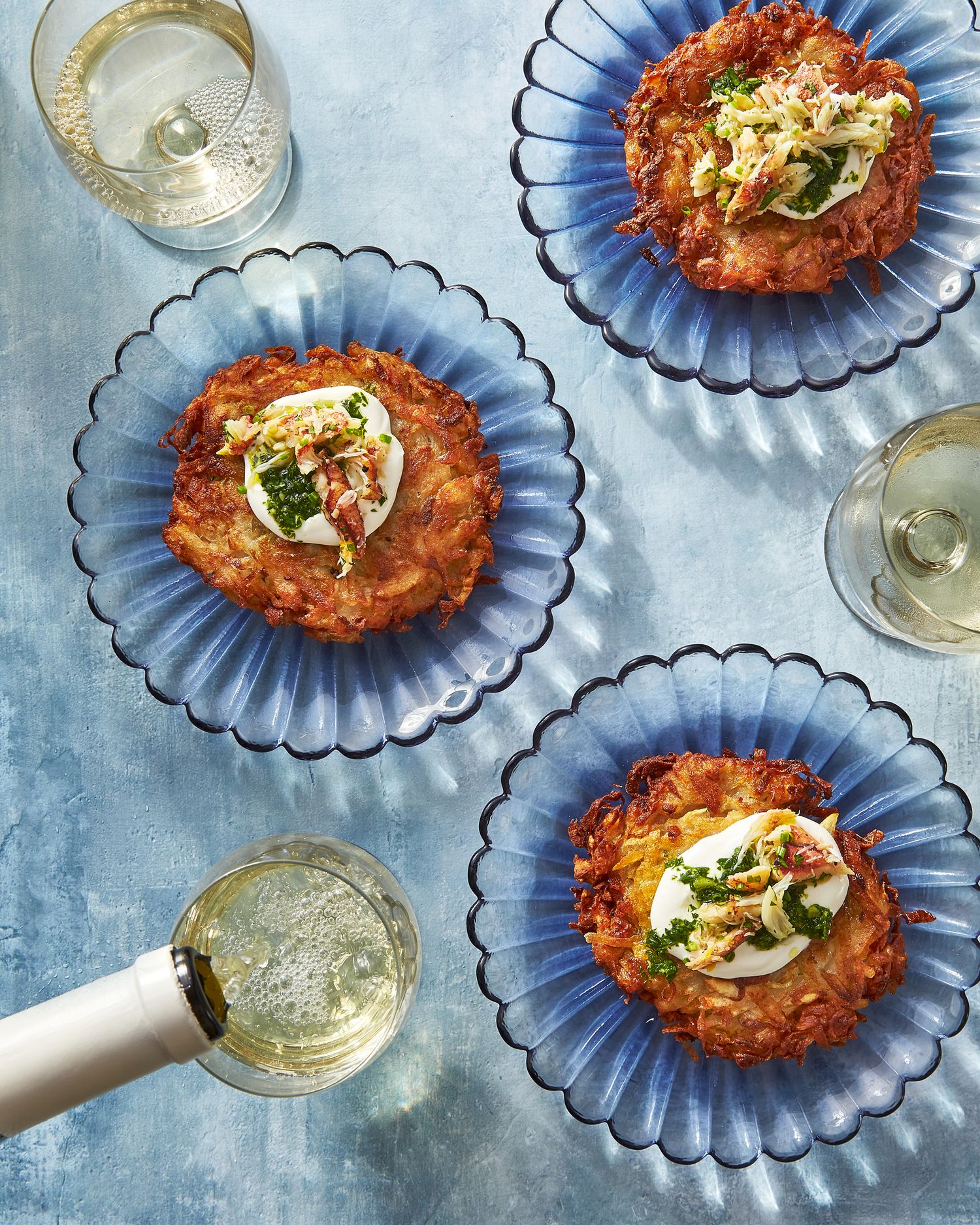 Crispy potato pancakes garnished with herbs on blue plates served with white wine.