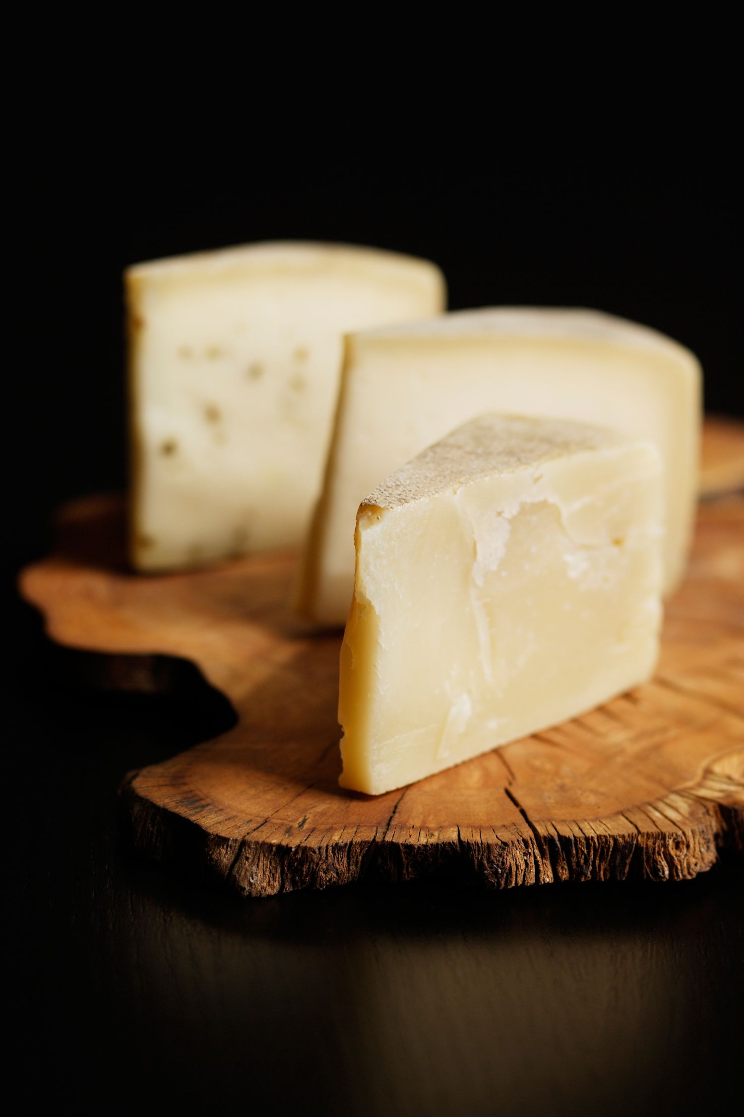 Two wedges of artisanal cheese with holes displayed on rustic wooden board against dark background.
