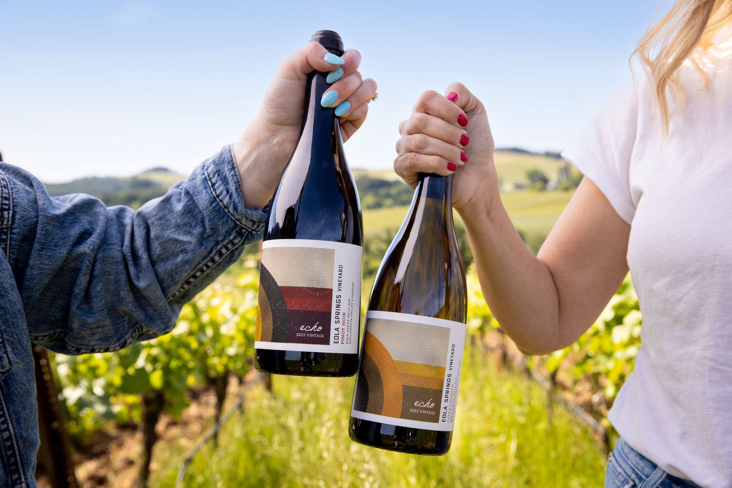 Two people toast with bottles of red wine in a sunny vineyard, one wearing a denim jacket and the other a white shirt with red nail polish.