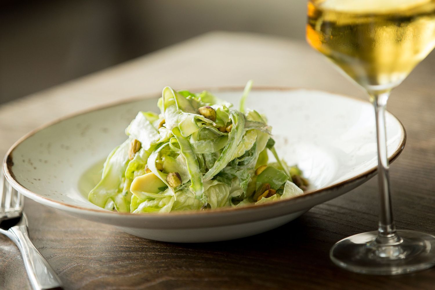Fresh green salad with shaved parmesan in a white bowl beside a glass of white wine.