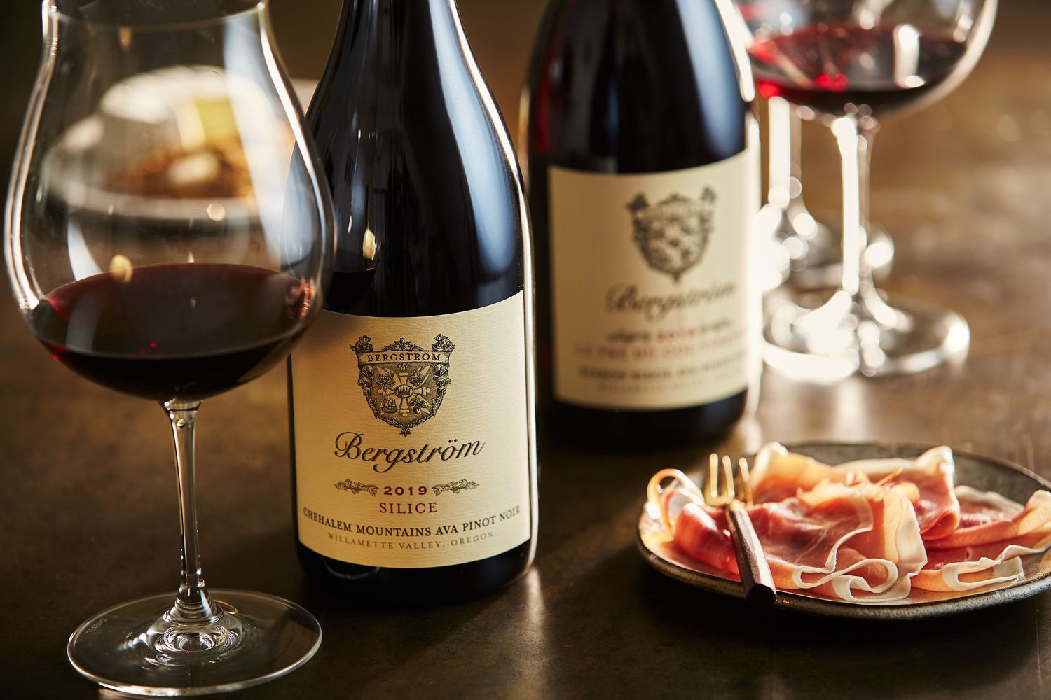 Wine bottles and glasses arranged with charcuterie on rustic wooden surface in warm lighting.