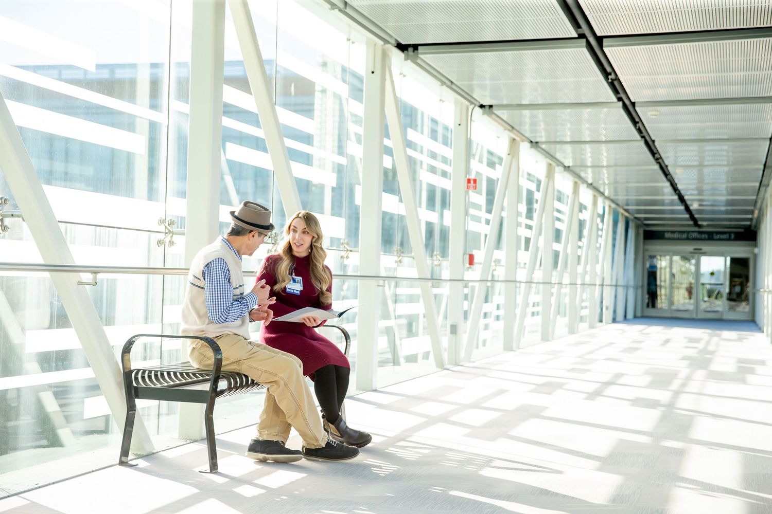Two people sitting together on bench in sunlit glass corridor.