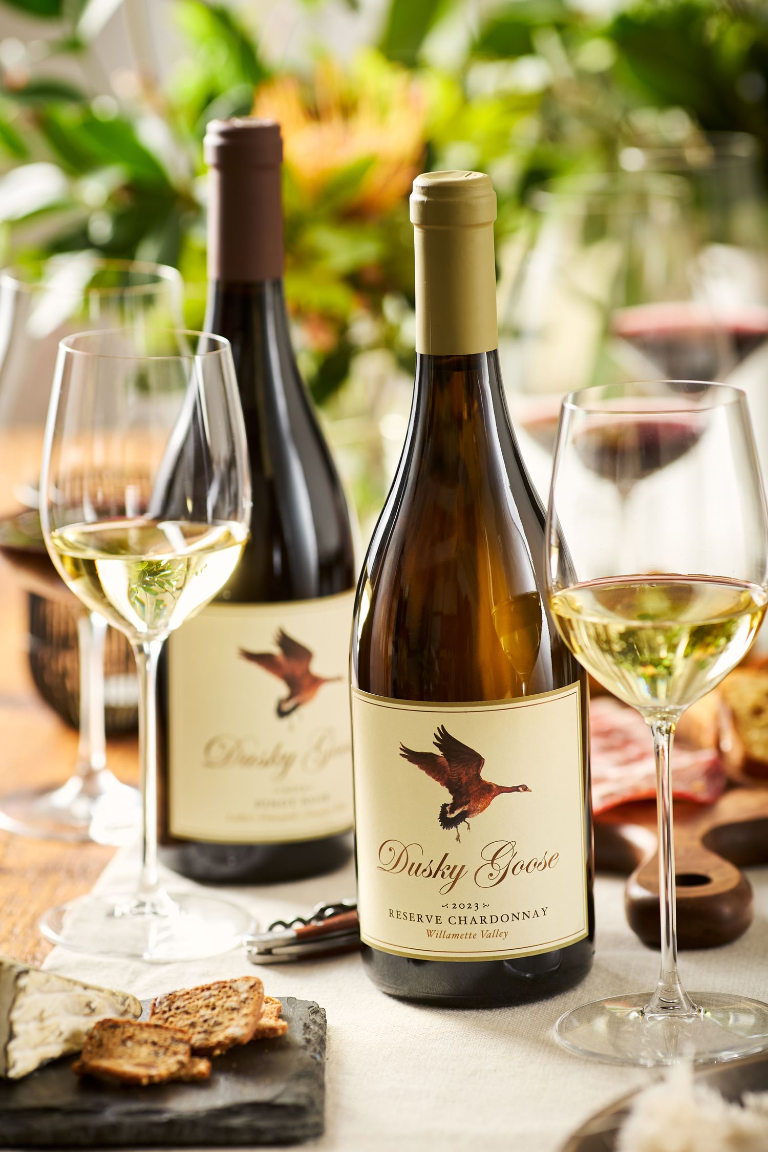Bottles of Ducky Goose wine with glasses of white wine and appetizers on wooden table.