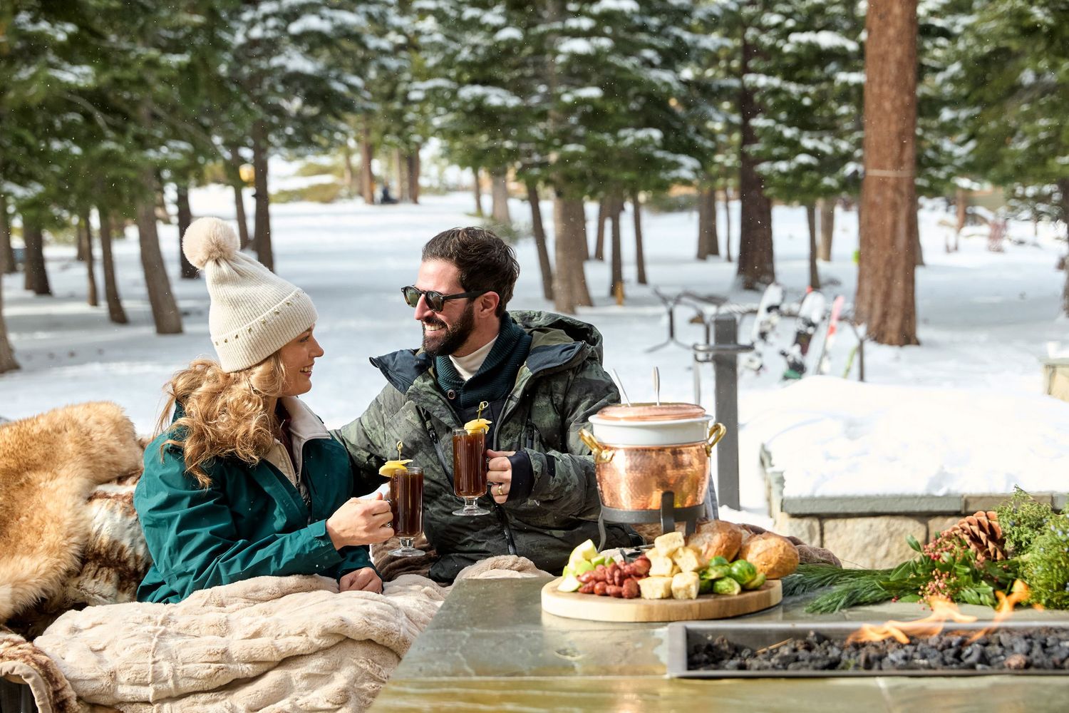 Two people sit together at an outdoor table enjoying coffee and conversation in a snowy winter forest setting.