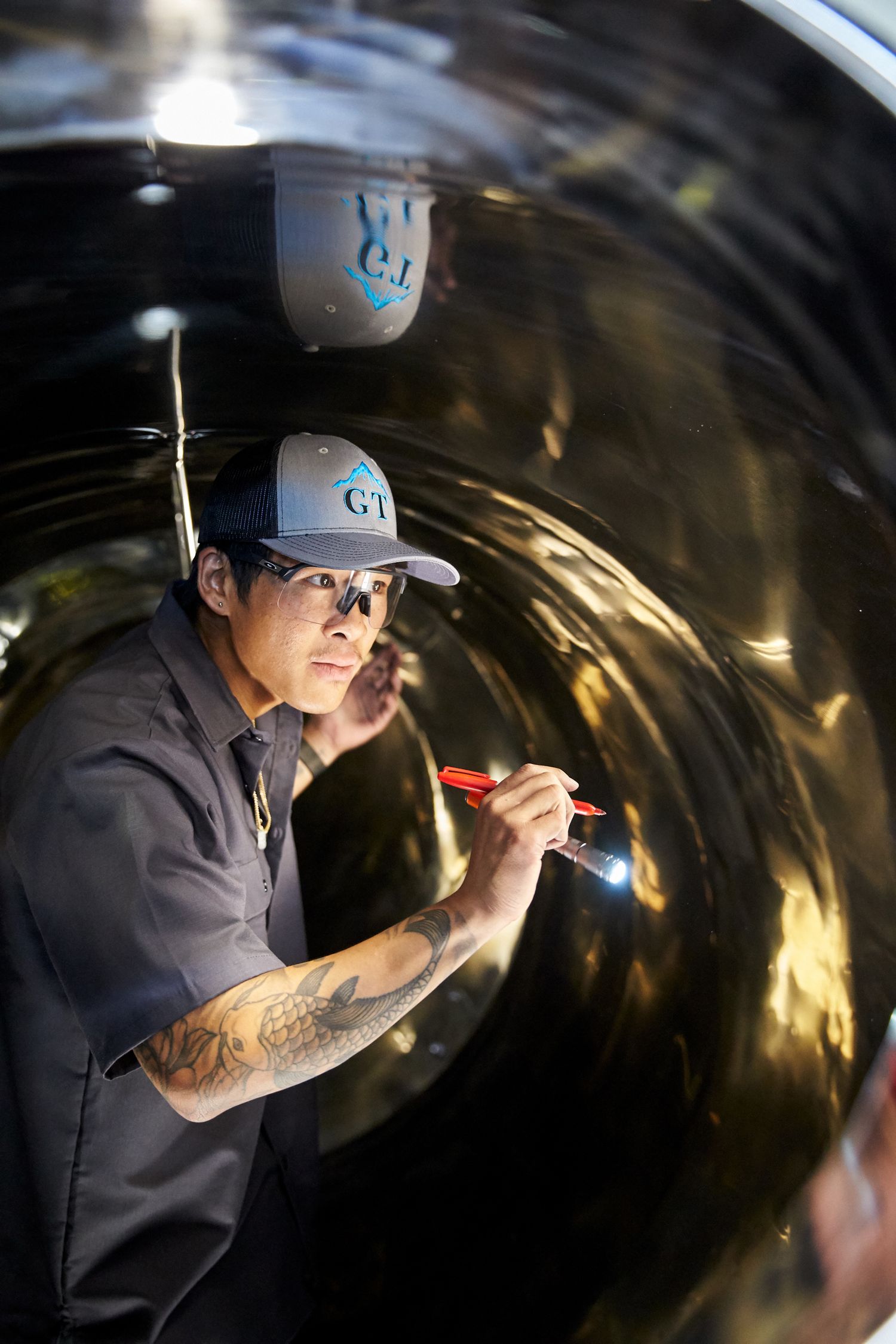 Worker in grey uniform and cap inspects interior of large industrial tank with flashlight.