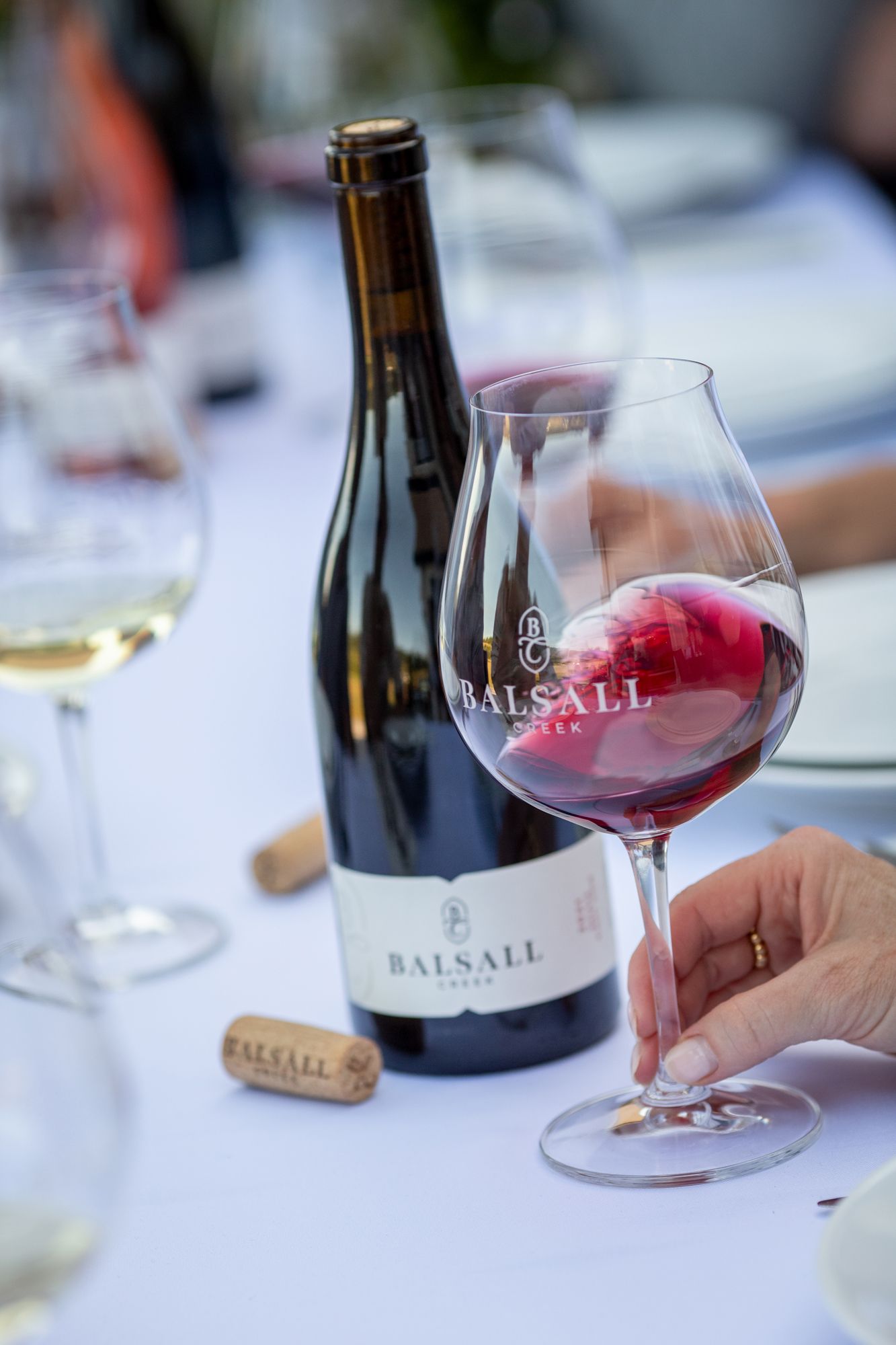 A glass of Balsall Creek red wine being swirled beside the bottle and cork on a white tablecloth.