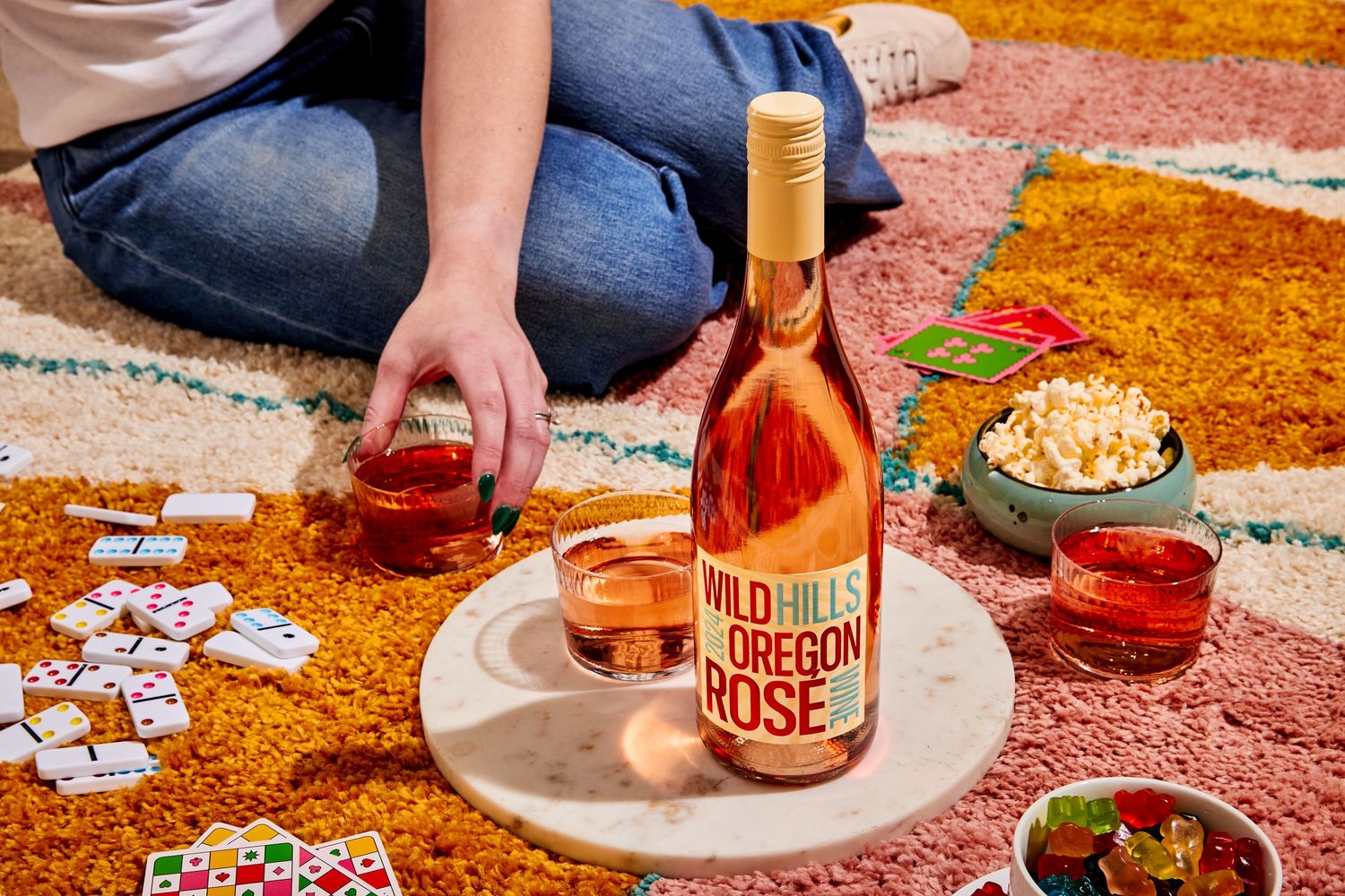Person sitting on a colorful rug playing dominoes beside a bottle of Wild Hills Oregon Rosé, filled glasses, and a bowl of popcorn.